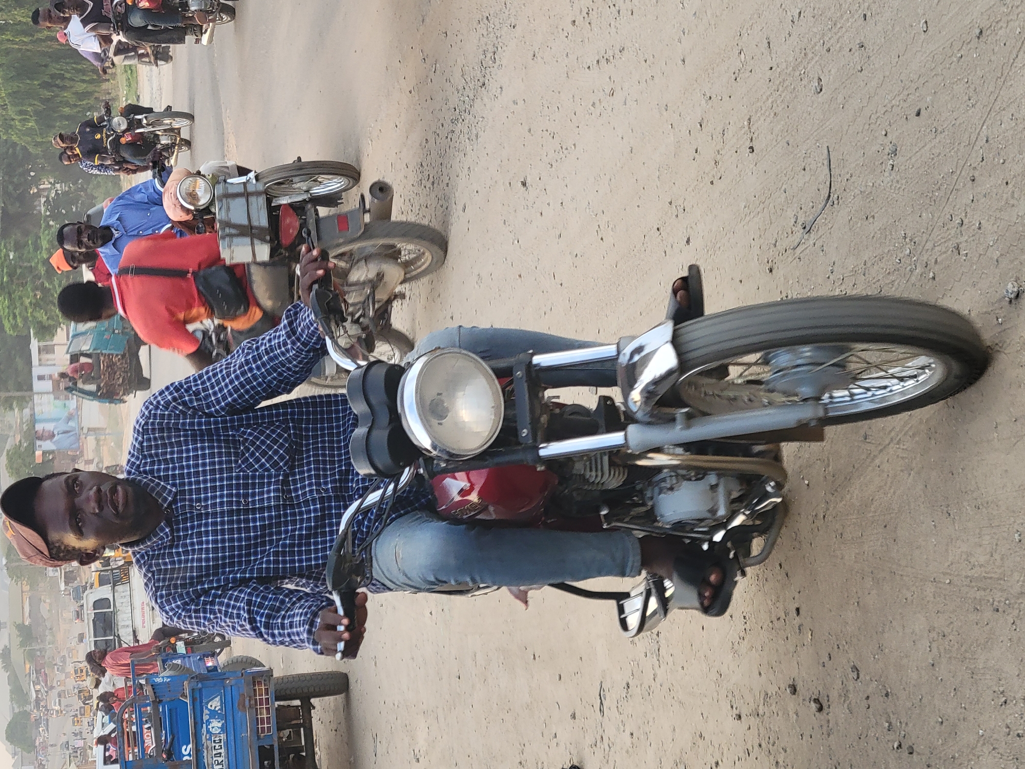 In Madalla, Nigeria, a man rides a red motorbike on a dusty, possibly unpaved road. He is wearing a blue checkered shirt, blue jeans, and sandals. He also wears a baseball cap and has a dark complexion. Other motorbikes and a blue truck can be seen on the road, suggesting traffic. The environment appears to be a typical city street with buildings and possibly trees in the background. The lighting suggests it is daytime.