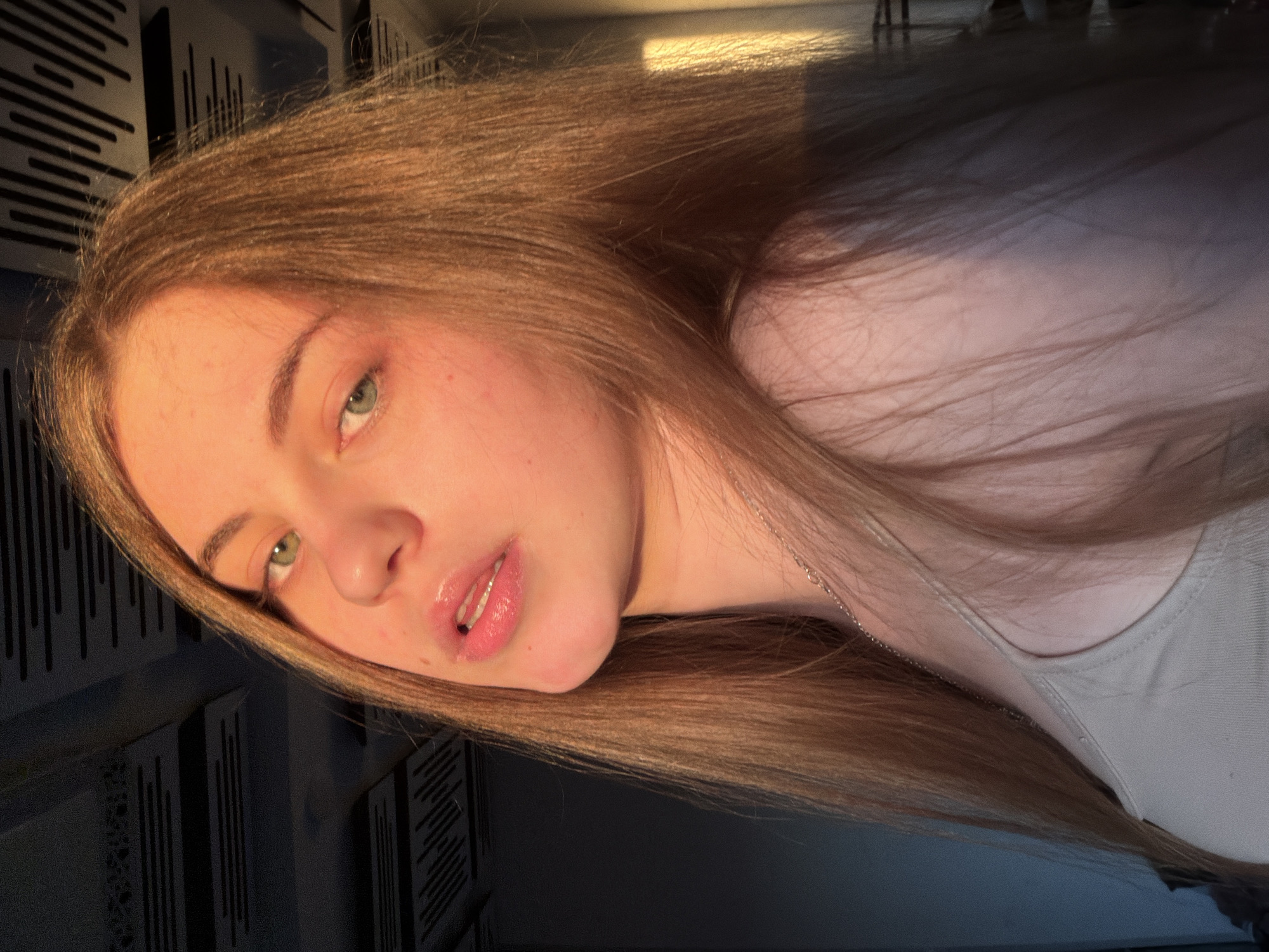 A young woman with long, light brown hair and green eyes is shown from the shoulders up, looking directly at the camera. She is wearing a light-colored tank top and a silver necklace. The lighting suggests it is either sunrise or sunset, casting a warm glow on her face and hair. The background is a dark ceiling with horizontal acoustic panels, and a wall with a vertical line of light. The overall impression is a portrait taken indoors. The location is unknown.