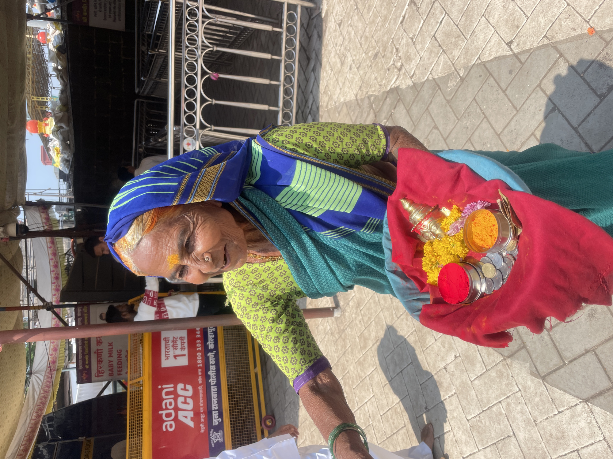 An elderly woman stands outdoors in Kolhapur, India, holding a blue basin draped with a red cloth. The basin contains small metal containers holding red vermilion powder and yellow turmeric powder, yellow marigold flowers, silver Indian rupee coins, and a small golden metallic deity figure. She is dressed in a green floral-patterned blouse, a blue and green sari, and has green bangles on her left wrist. A silver nose ring is visible, and a yellow mark adorns her forehead. She smiles subtly, looking towards the left of the frame.
The background features a public area with a prominent red barrier displaying "Adani ACC" branding, Marathi text, and "Juna Rajwada Police Thana, Kolhapur." A sign for "Baby Milk Feeding" is also visible. Metallic crowd control barriers, black portable structures, and an overhead fabric canopy define the space. Behind these, parts of a structure with orange and red conical tops, characteristic of temple architecture, are discernible. The ground is paved with interlocking concrete blocks, and the scene is illuminated by bright daylight, casting shadows. Several other individuals are present in the background, partially obscured or out of focus, including a man in a white shirt near the red barrier. A security camera is mounted overhead.