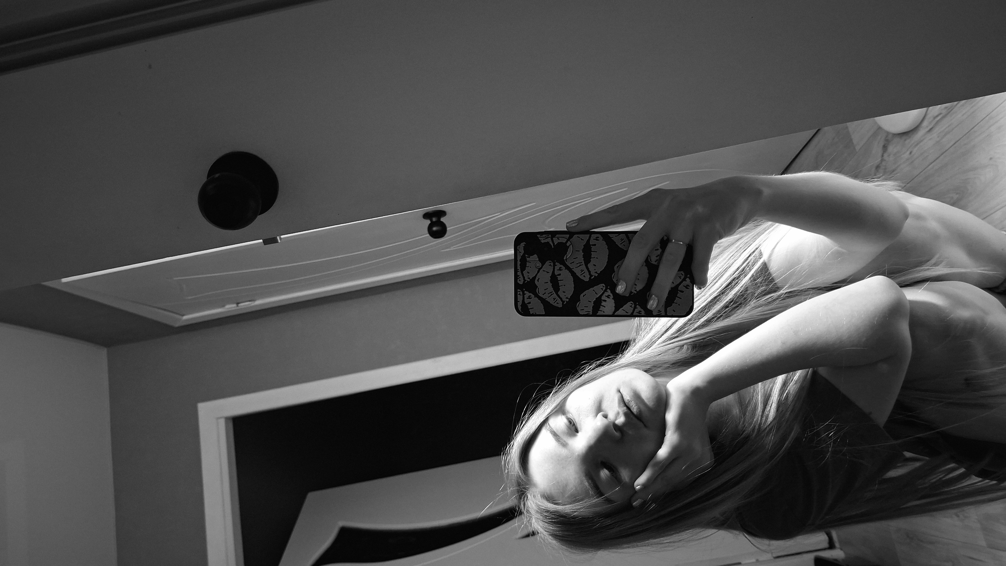 The image is a black and white photograph taken in what appears to be an indoor setting, possibly a room. A woman with long hair is visible, seemingly holding a cell phone in her hand, perhaps taking a selfie. The phone has a patterned case with lip imprints. The woman is positioned in a way that her face is partially obscured by her hand and hair.
In the background, a door is visible, along with a door knob. A portion of the room's architecture, including the ceiling and walls, can be seen.
There is no specific information visible in the image that confirms the location to be Chaikovsky, Russia.