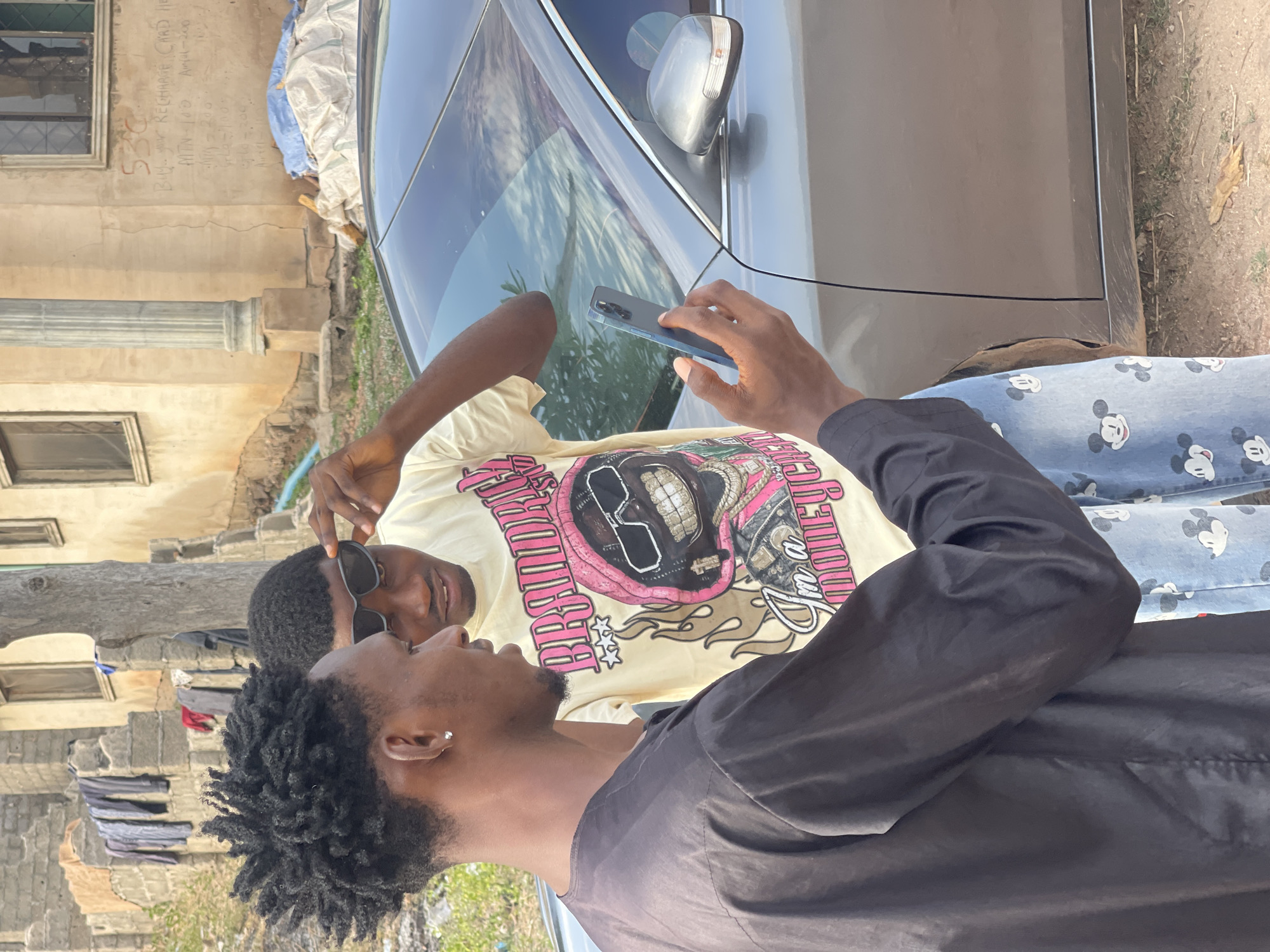Two young men are standing next to a silver car in an outdoor setting. One of the men is holding a smartphone in his hand, pointing the camera upwards. He is wearing a black shirt and Mickey Mouse print jeans. The other man is wearing a light yellow t-shirt with a pink and black graphic design and sunglasses. The background shows buildings, one of which has graffiti or writing on the wall, possibly advertisements for recharge cards. The location could be Lokoja, Nigeria, given the information. The subjects are interacting with each other while near the car, possibly taking a picture or video.