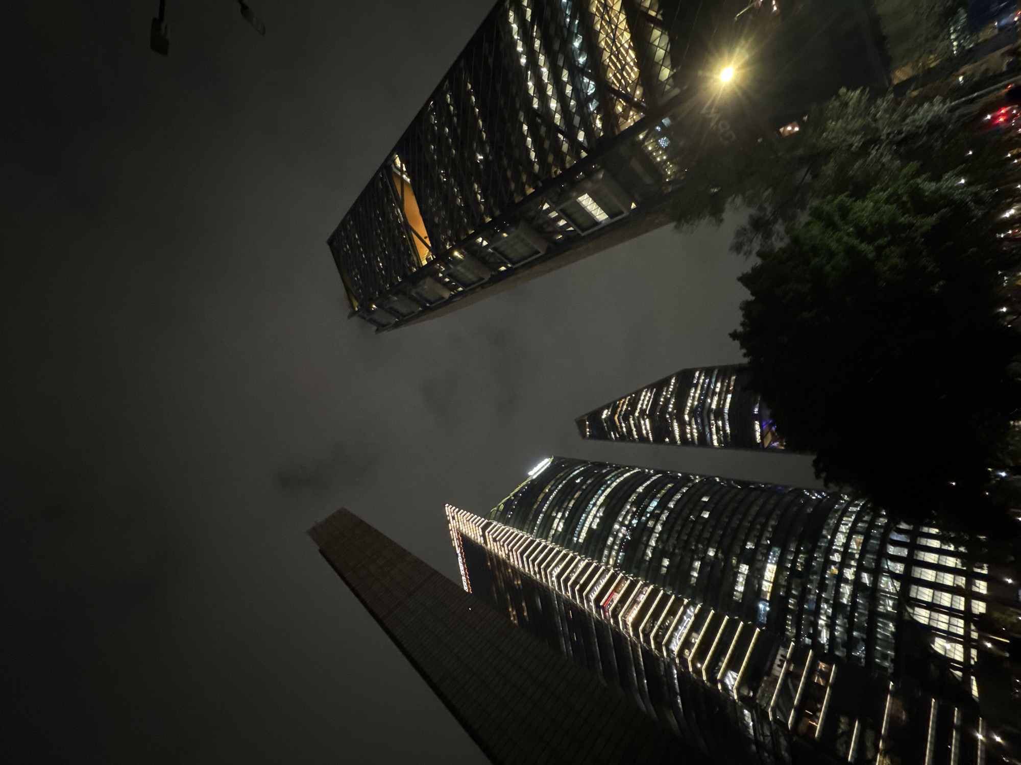 The image presents a dramatic low-angle view of multiple modern skyscrapers against a dark, overcast night sky. The perspective looks sharply upwards, emphasizing the immense height and contemporary design of the urban architecture.
In the upper left quadrant, a large, rectangular skyscraper stretches diagonally across the frame. It features a grid-like facade with numerous brightly lit windows and internal illumination, creating striking patterns of light and shadow. Below and towards the right, a cluster of additional skyscrapers rises. One prominent building exhibits a distinctive curved or rounded facade, adorned with parallel strips of external lighting that emit a warm glow. Adjacent to it, a narrower, pointed skyscraper also showcases internally lit windows. The dark tones of the building exteriors allow the vivid internal and external lights to stand out prominently.
The sky is heavily overcast, appearing dark gray and hazy, suggesting either low clouds or fog that partially obscure the upper sections of the structures and diffuse the light sources. A bright, starburst-like streetlight is visible in the upper right, partially obscured by the dark foliage of trees that extend into the frame from the right edge. The scene depicts a bustling, modern metropolis at night, with the impressive architecture dominating the visual space. No specific activities or people are visible, and there is no discernible text.