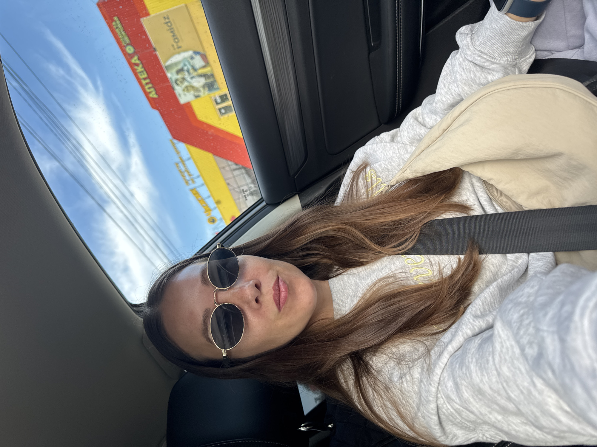A woman with long brown hair is sitting in the passenger seat of a car. She is wearing oval sunglasses, a grey sweatshirt, and a light-colored shoulder bag. Her seatbelt is fastened. Outside the car window, a brightly colored building with the text "АПТЕКА" (Pharmacy) and "Famidz" is visible against a blue sky with some clouds. The building appears to be a commercial establishment in the city of Chaikovsky, Russia.