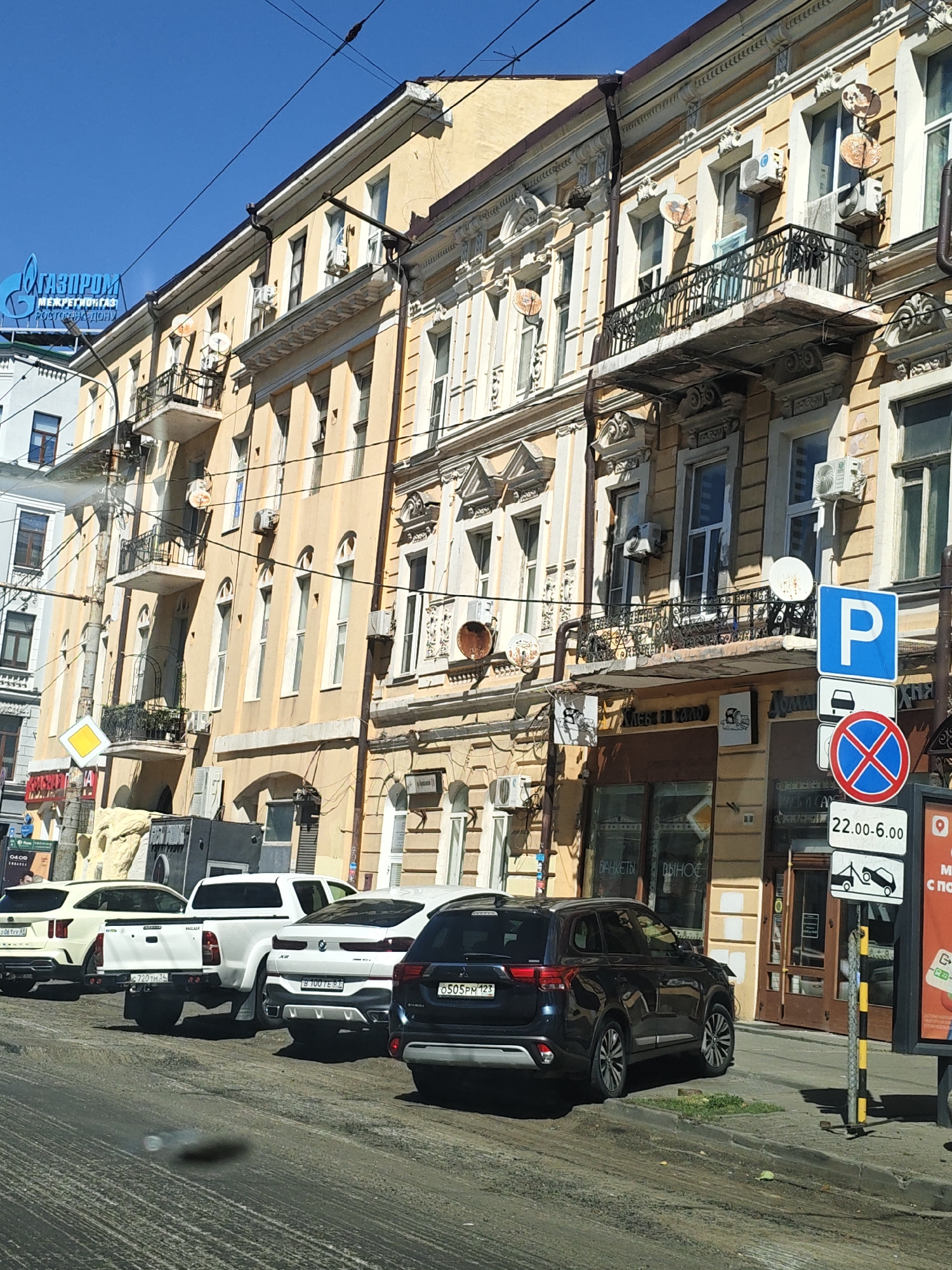 The image depicts a street scene in Rostov-on-Don, Russia, under a clear blue sky. The foreground is dominated by a dirty, uneven road surface. Parked along the right side of the road are several cars, including a dark SUV, a white BMW X6, a white Toyota Hilux pickup truck, and a light-colored SUV.
The background features an old, multi-story building with ornate architecture, painted in a pale yellow hue. The building has numerous windows, some with balconies adorned with wrought-iron railings. Air conditioning units and satellite dishes are visible attached to the building's facade. A large blue sign with the Gazprom logo and text is mounted on the building.
Various street signs are present, including a blue parking sign, a red circle with a diagonal cross, and a sign indicating parking hours from 22:00 to 6:00. A tow truck symbol is also visible on a sign. Some storefronts are visible at the ground level of the building. The overall impression is a sunny day in an urban environment with older architecture.