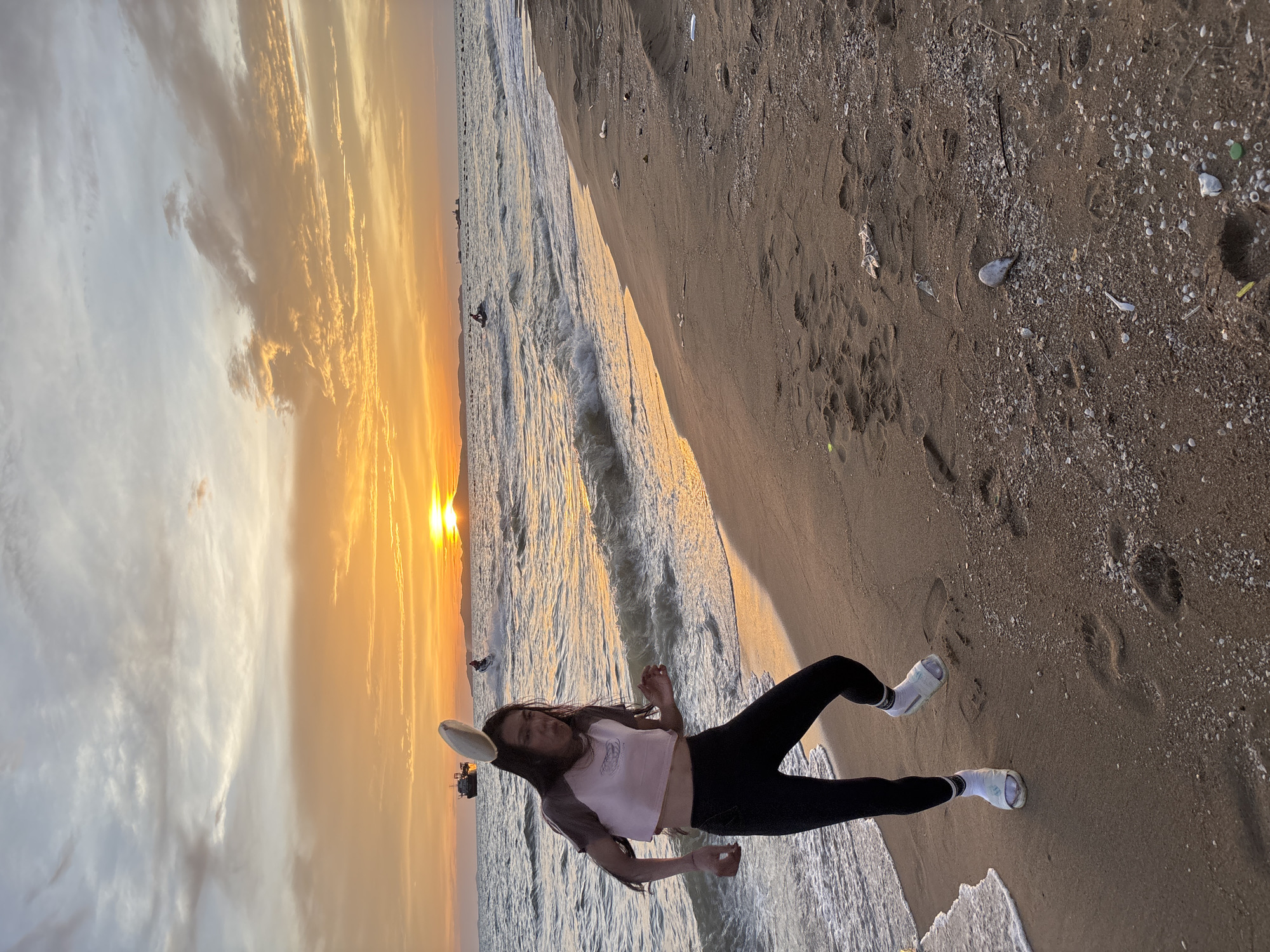 A person stands on a sandy beach in Pattaya City, Thailand, with the ocean and a sunset in the background. The person is wearing a light pink cropped t-shirt, black leggings, white socks, and white slides. A light-colored frisbee is balanced on their head. The person's facial expression suggests concentration or perhaps a playful moment. The ocean has small waves rolling onto the shore, and the sky is filled with warm colors from the setting sun. Several boats and jet skis are visible on the water in the distance. The beach itself is sandy with some small shells and debris scattered on the surface. The overall scene captures a moment of leisure and activity at the beach during sunset.