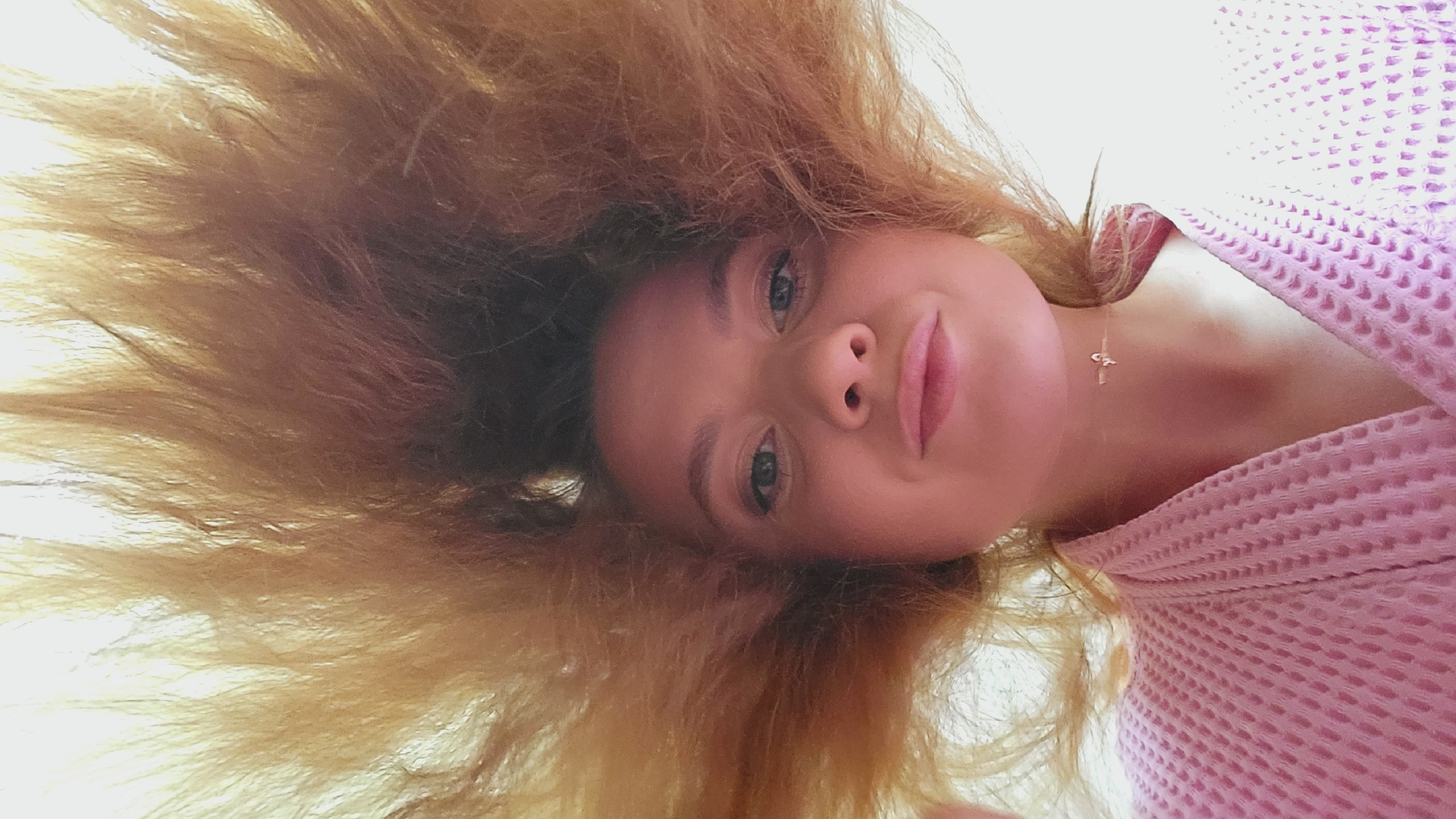 A selfie of a woman lying on her back is presented from an upward angle. Her hair, primarily light brown, surrounds her face. She has blue eyes and pink lips. She is wearing a light pink textured garment, and a gold necklace with a cross pendant is visible. No location is depicted.