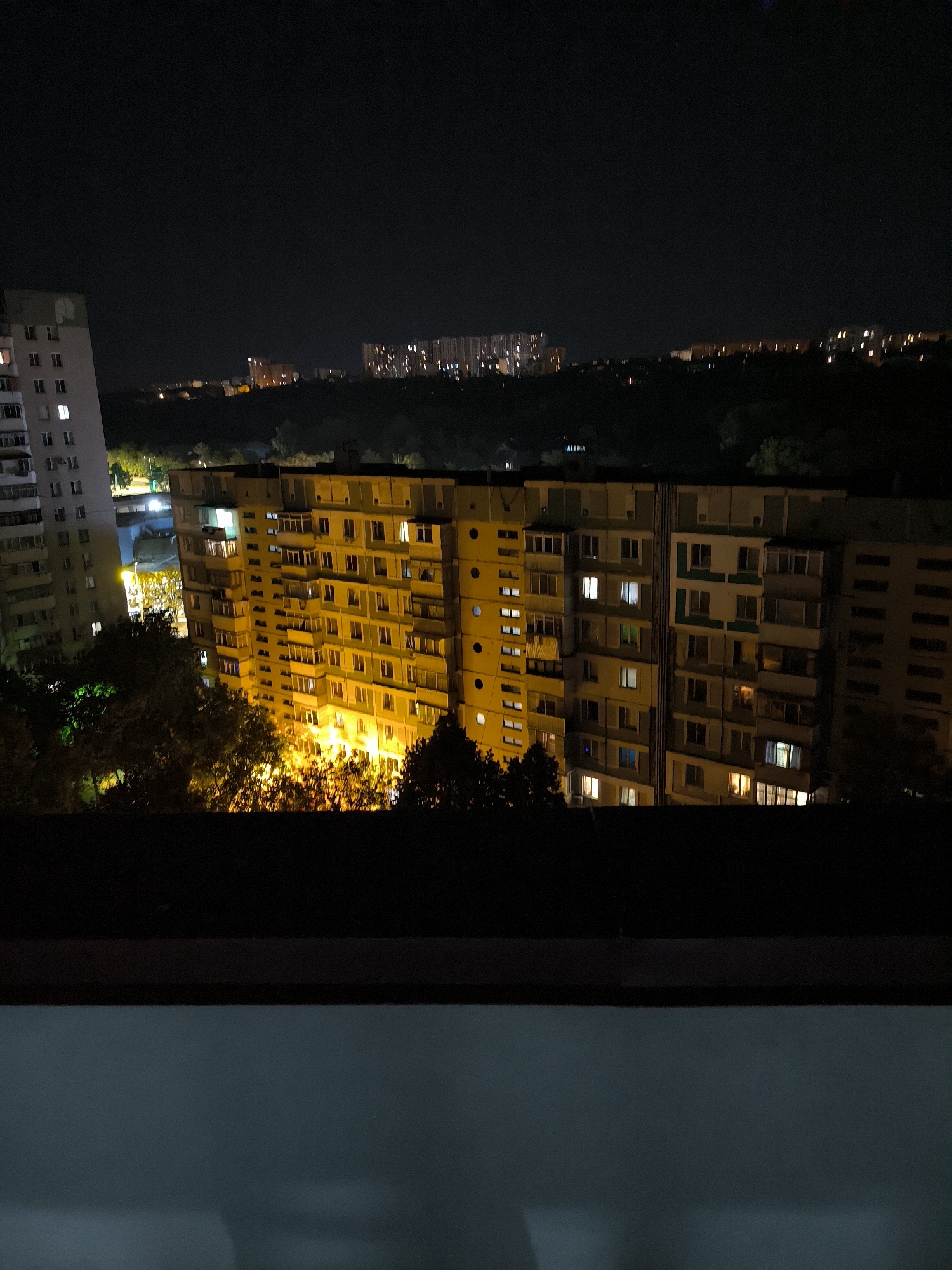 The image depicts a nighttime view of a residential area in Chișinău, Moldova. The foreground features a dark, blurred balcony railing, suggesting the photo was taken from an elevated position. Dominating the mid-ground is a large, multi-story apartment building illuminated by numerous lit windows, indicating occupancy. To the left, another apartment building, taller and lighter in color, also shows lit windows. Beyond these buildings, a line of trees is visible, their leaves illuminated by warm, artificial light from below, creating a fiery effect. In the distance, more apartment buildings and the hazy outlines of distant structures can be seen against the dark sky. The overall scene conveys a typical urban environment at night, with artificial lighting casting a glow on the buildings and surrounding landscape.