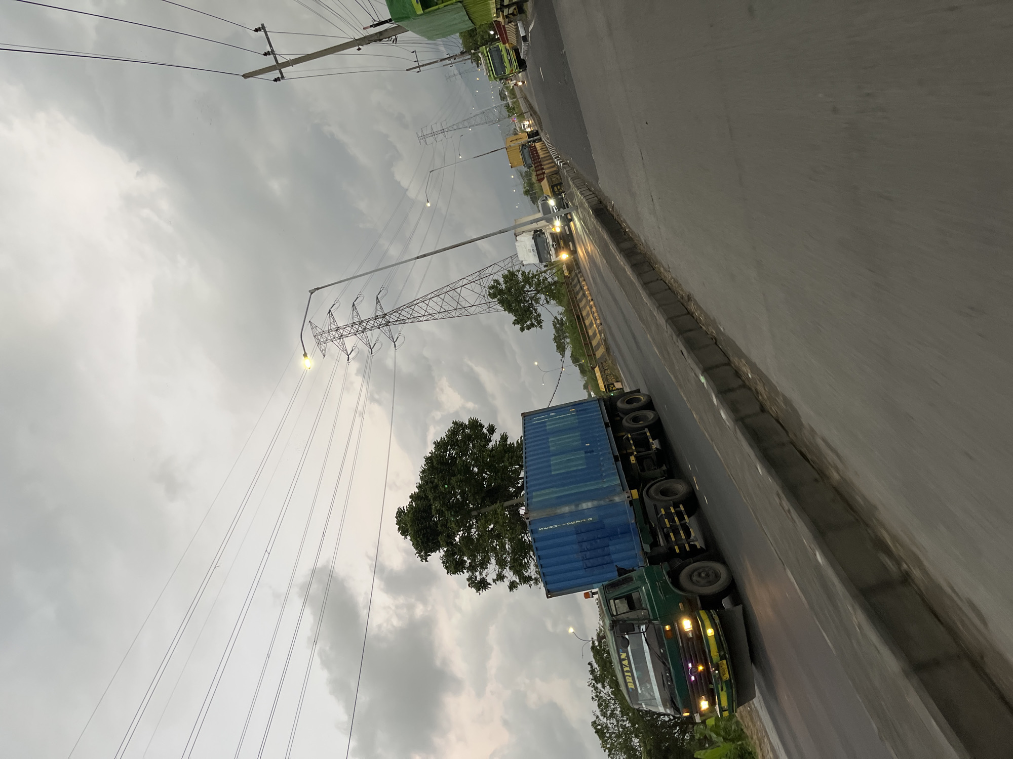 The image shows a truck with a blue shipping container on a road, likely in Pamotan, Indonesia. The truck is dark green and has "BRIYANI" written on its side. The container is predominantly blue with some white text and markings. The truck is positioned on the right side of the road, and the image is oriented vertically.
The scene appears to be a road with multiple lanes, and other vehicles are visible in the distance, suggesting traffic. Tall power lines and utility poles are prominent features of the landscape, stretching across the sky. The weather appears overcast or cloudy, with a muted light, indicating it might be dawn, dusk, or a heavily clouded day. The road surface is wet, hinting at recent rain or humidity.
The overall impression is that of a transportation scene in an Indonesian setting, possibly during a time of day with less direct sunlight. The image's composition, with the truck and power lines dominating the frame, creates a sense of scale and the industrial or logistical nature of the scene. There are no people clearly visible in the image. The focus is on the truck, the container, the infrastructure, and the atmosphere.