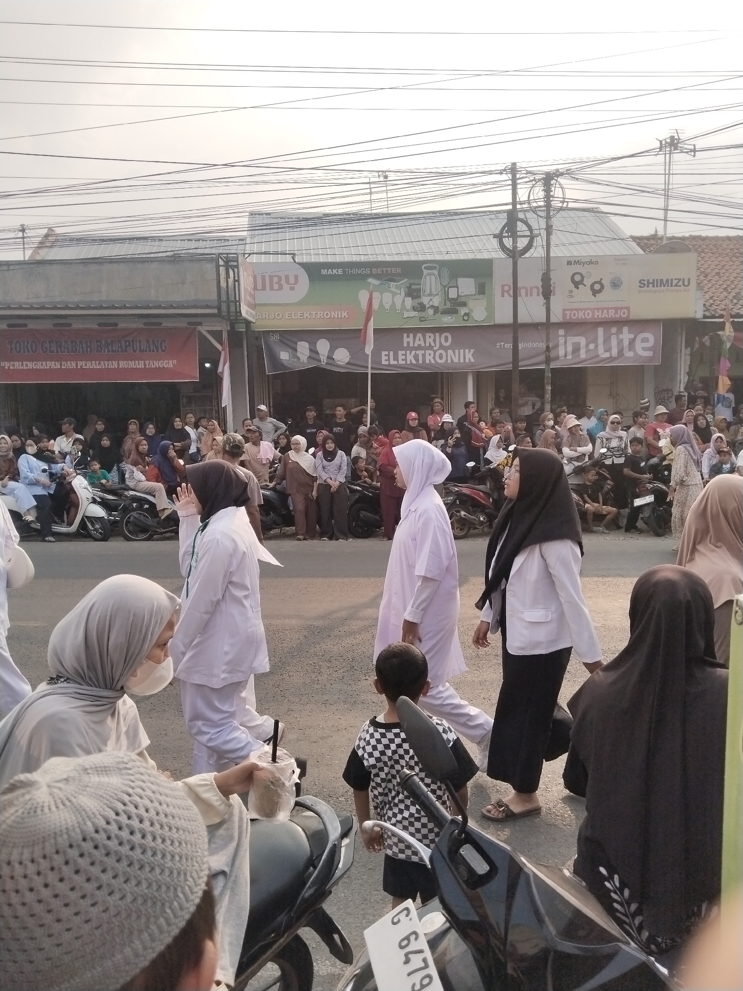 The image depicts a street scene in Balapulang Kulon, Indonesia, possibly during a procession or gathering. A crowd of people, many wearing headscarves and traditional clothing, are visible on the right side of the frame, lining the street. Several motorcycles are parked along the roadside. In the foreground, a child in a checkered shirt walks past. Three women in white medical uniforms are walking from left to right across the street, with one carrying a drink. Behind them, more people are gathered. Buildings with signs in Indonesian, including "TOKO GERABAH BALAPULANG" and "HARJO ELEKTRONIK," are visible in the background. The sky is overcast.