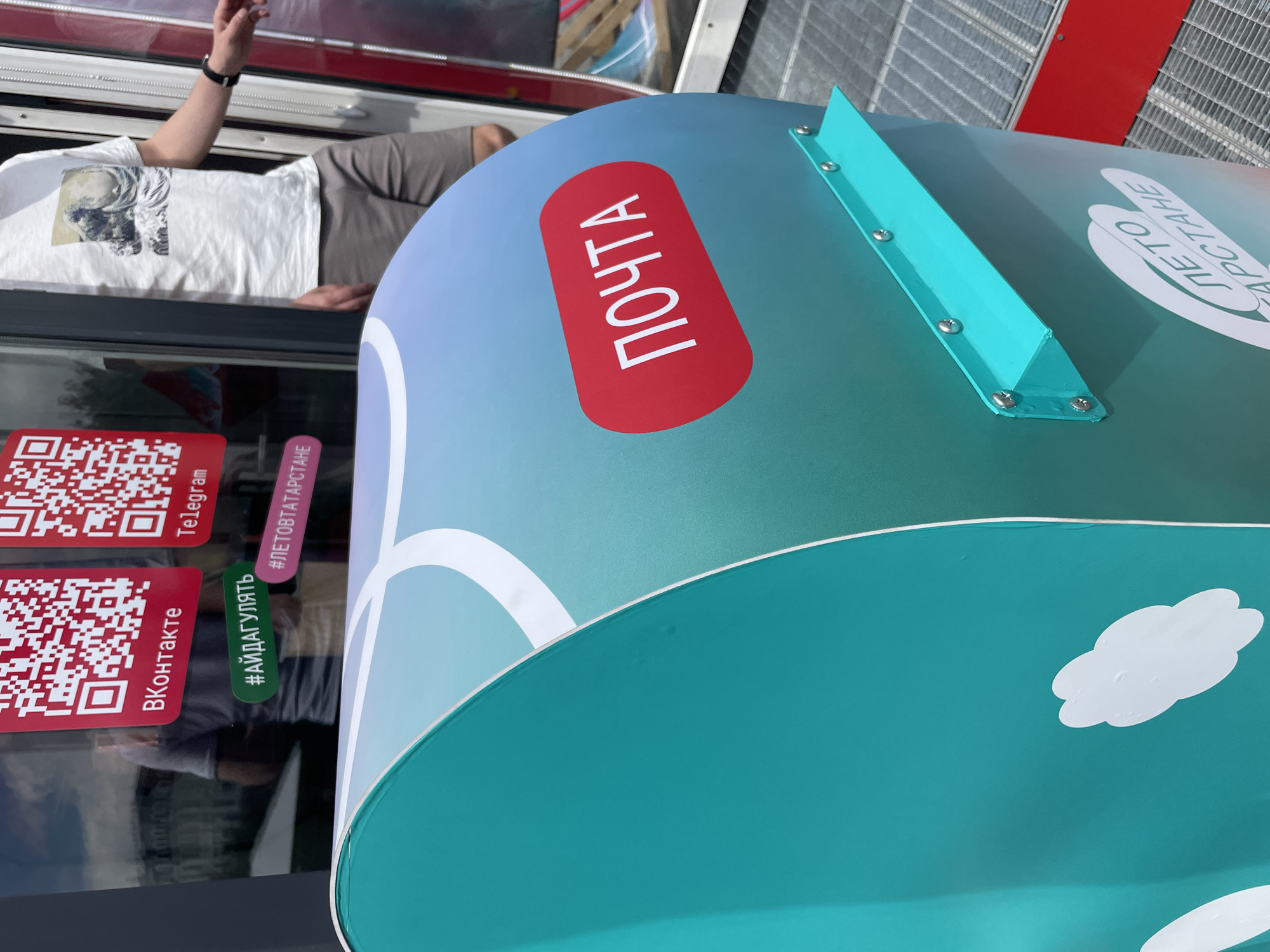 A close-up, eye-level shot features a teal and blue gradient-colored postbox in Kazan, Russia. The postbox has a red oval sticker with the white text "ПОЧТА" (POST). Above the postbox, two red QR codes are visible on a glass surface, labeled "ВКонтакте" (VKontakte) and "Telegram." Below the QR codes are hashtags "#АЙДАГУЛЯТЬ" (LET'S GO FOR A WALK) and "#ЛЕТОВТАТАРСТАНЕ" (SUMMER IN TATARSTAN). A person in a white t-shirt with a Japanese wave design and gray shorts is partially visible to the right, standing near a doorway. The postbox also has white cloud-like graphics and a smaller teal shelf attached with screws. The background includes a metal grating floor and red accents.