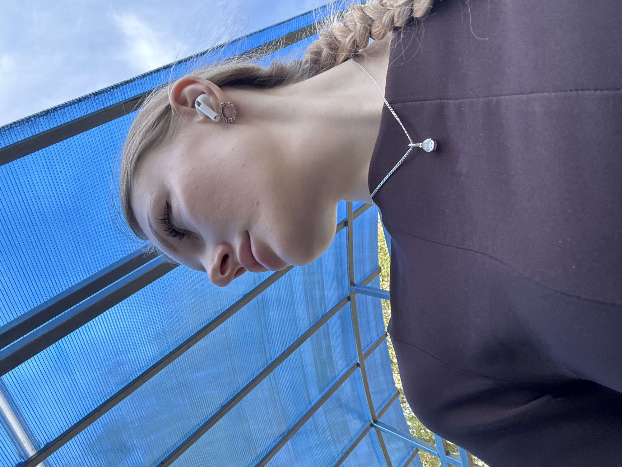 A woman is viewed from a low angle beneath a blue, corrugated polycarbonate roof. Her face, in profile, shows light skin, blond hair in a braid, and an earbud. A silver necklace with a crystal pendant is visible against her dark clothing. The location is likely in Perm, Russia, based on other information.