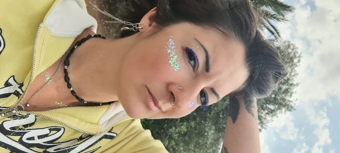 An outdoor, eye-level portrait shot in Mate Asher Regional Council, Israel, features a female individual looking directly at the camera with a contemplative expression. Her dark hair is styled in an updo, with strands framing her face. She has dark eyebrows and her eyes are accentuated with bright blue eyeliner/mascara. Glitter is applied to her left cheekbone and near her right eye. Her nose has a small stud piercing on the right nostril and a hoop piercing on the septum. She wears multiple earrings in her left ear, including a dangle earring with a crescent moon charm and chains. Around her neck, she has a black beaded choker and a longer silver chain with multiple small pendants. She is dressed in a yellow zippered hoodie, partially revealing a white hood lining. A black tattoo is visible on her upper left arm/shoulder. The background is blurred, showing green foliage, including palm fronds on the left and a deciduous tree, under a partly cloudy sky, indicating daylight.