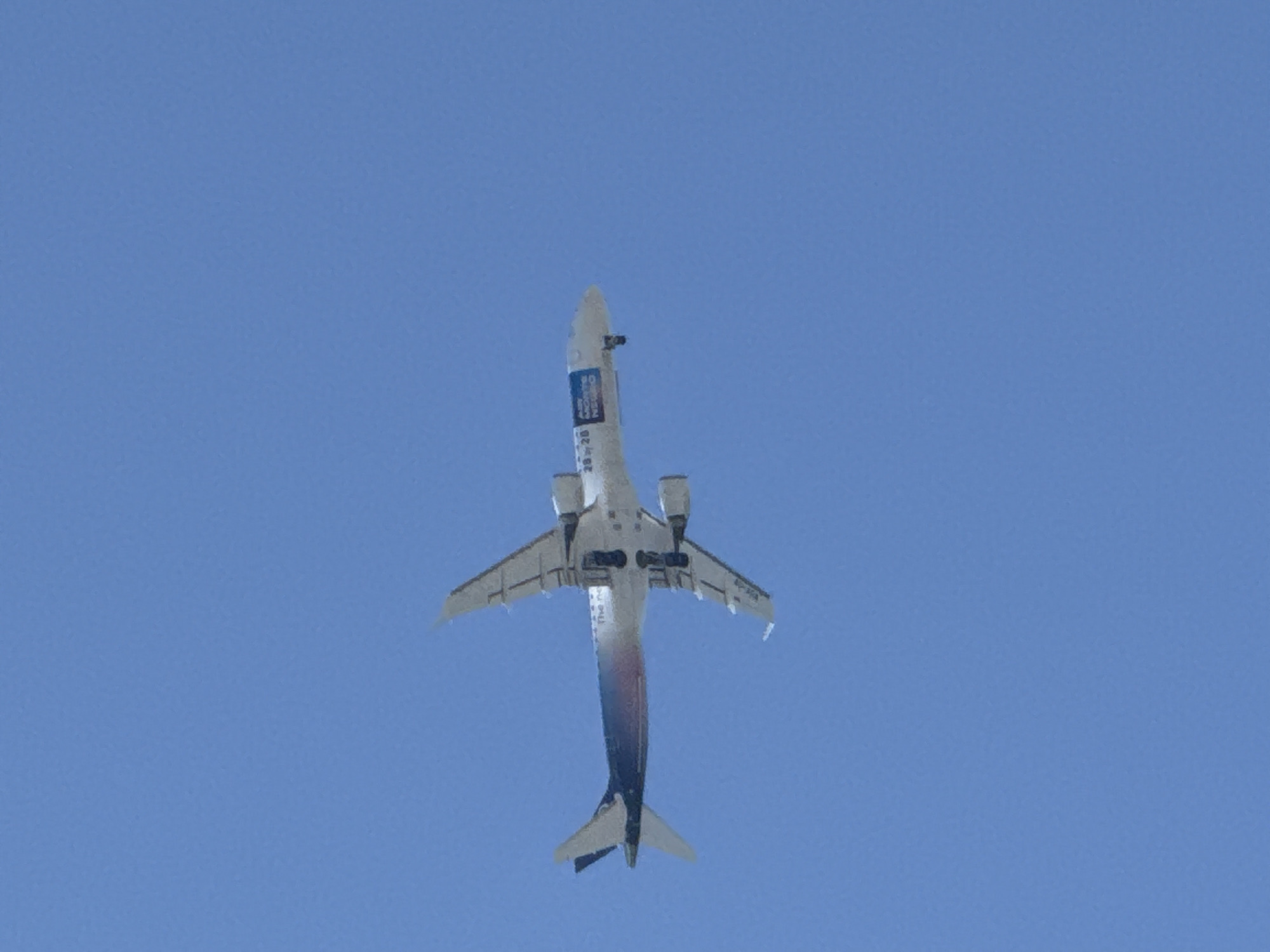 An airplane is captured mid-flight against a clear blue sky. The aircraft appears to be an Airbus A320, painted in a white livery with blue and gradient accents towards the tail. Visible text on the fuselage includes "The new" and a logo with "ASKY MORNING NEGRO" in blue and white. The plane is angled with its underside visible, showing its landing gear extended. The perspective suggests the photo was taken from below, looking up. There are no other discernible objects or people in the frame. The location in Budva, Montenegro, is not evident from the image.