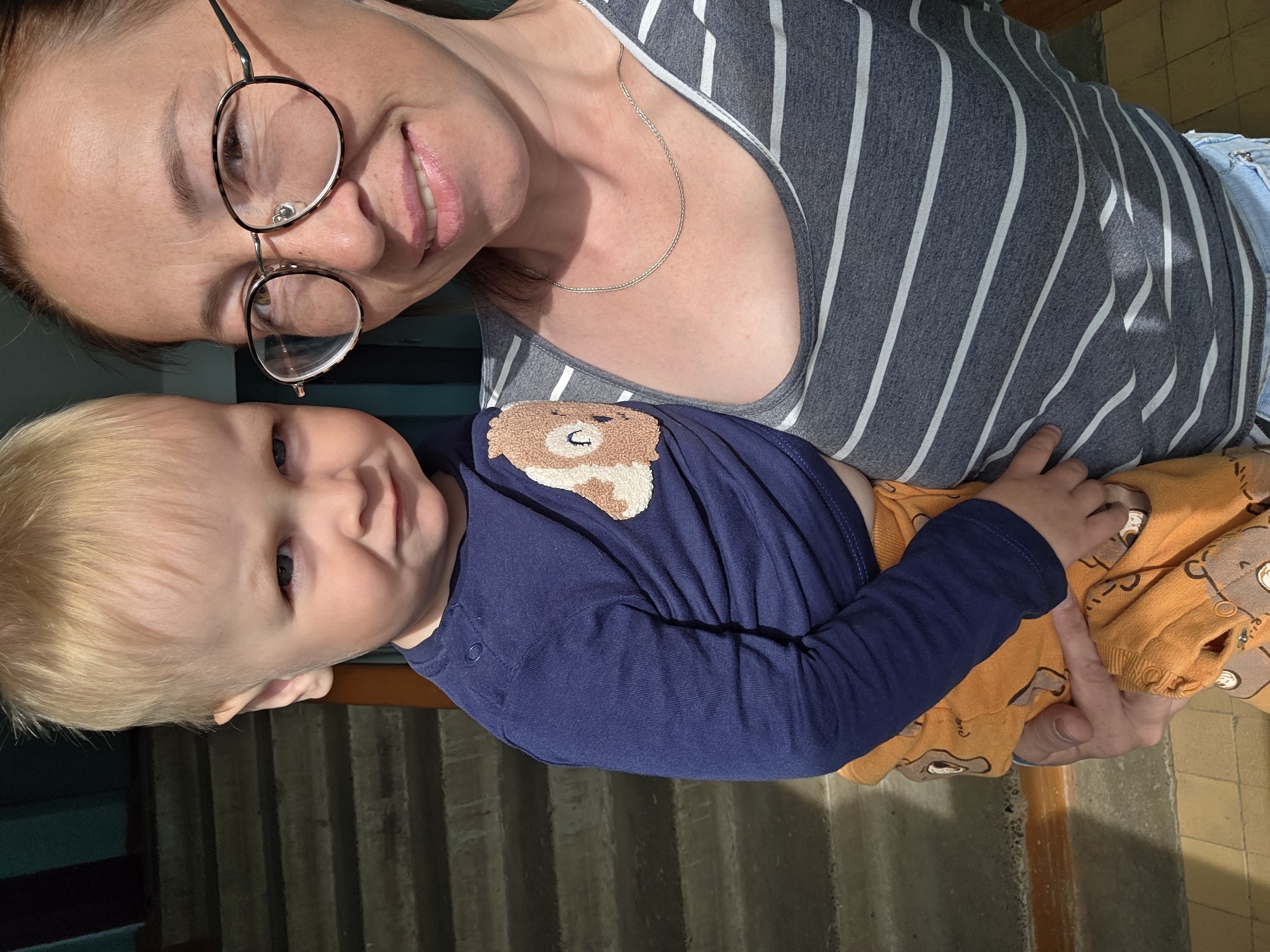 A medium shot shows a fair-skinned woman with dark brown hair, wearing round glasses and a gray and white striped tank top, holding a blond-haired toddler. The woman is smiling and looking at the camera, while the toddler is looking away with a displeased expression. The toddler is wearing a navy blue long-sleeved shirt with a bear graphic and orange pants with a bear pattern. The background shows a staircase and a wall. The overall scene appears to be indoors. The location is the city of Chaikovsky, Russia.