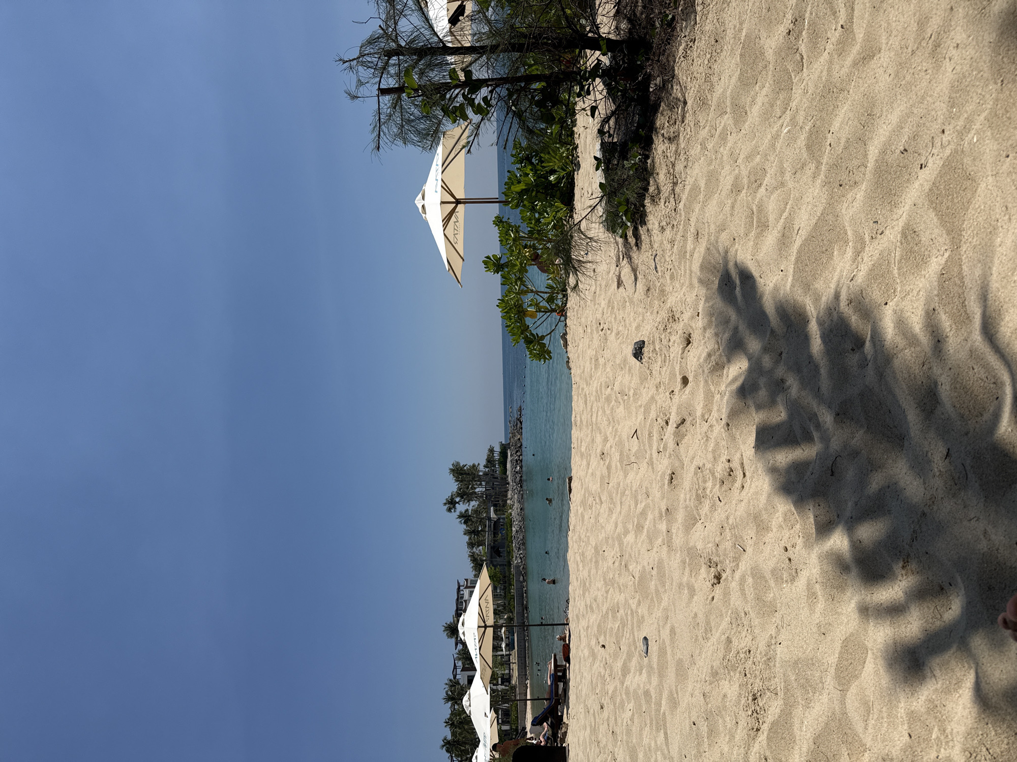 The scene depicts a beachfront environment under a clear blue sky in Nha Trang, Vietnam. The foreground consists of light tan sand, exhibiting ripples and partially covered by the dark, elongated shadows of unseen overhead foliage. The tip of a human foot, specifically toes, is visible at the bottom center, suggesting a low vantage point from the sand.
In the midground, towards the left, two white hexagonal beach umbrellas are open, both displaying the word "SATINA" in red lettering. Beneath the leftmost umbrella, a person is seated on a dark lounge chair facing the water. Another individual is partially obscured behind the second umbrella. The turquoise ocean water extends from the midground towards the horizon. Approximately 6-8 individuals are visible swimming or wading in the clear water. A dark object, possibly a rock or shell, rests on the sand to the left of the umbrellas.
Further to the right in the midground, a third "SATINA" umbrella is situated behind dense green bushes. Adjacent to these bushes, a tall, partially defoliated tree with thin branches stands prominently.
The background on the left features a rocky shoreline barrier leading to a cluster of multi-story buildings, consistent with resort architecture, nestled among numerous palm trees. The horizon line across the ocean is distinct and unbroken, meeting the clear sky. Buoys or markers are visible in the distant water.