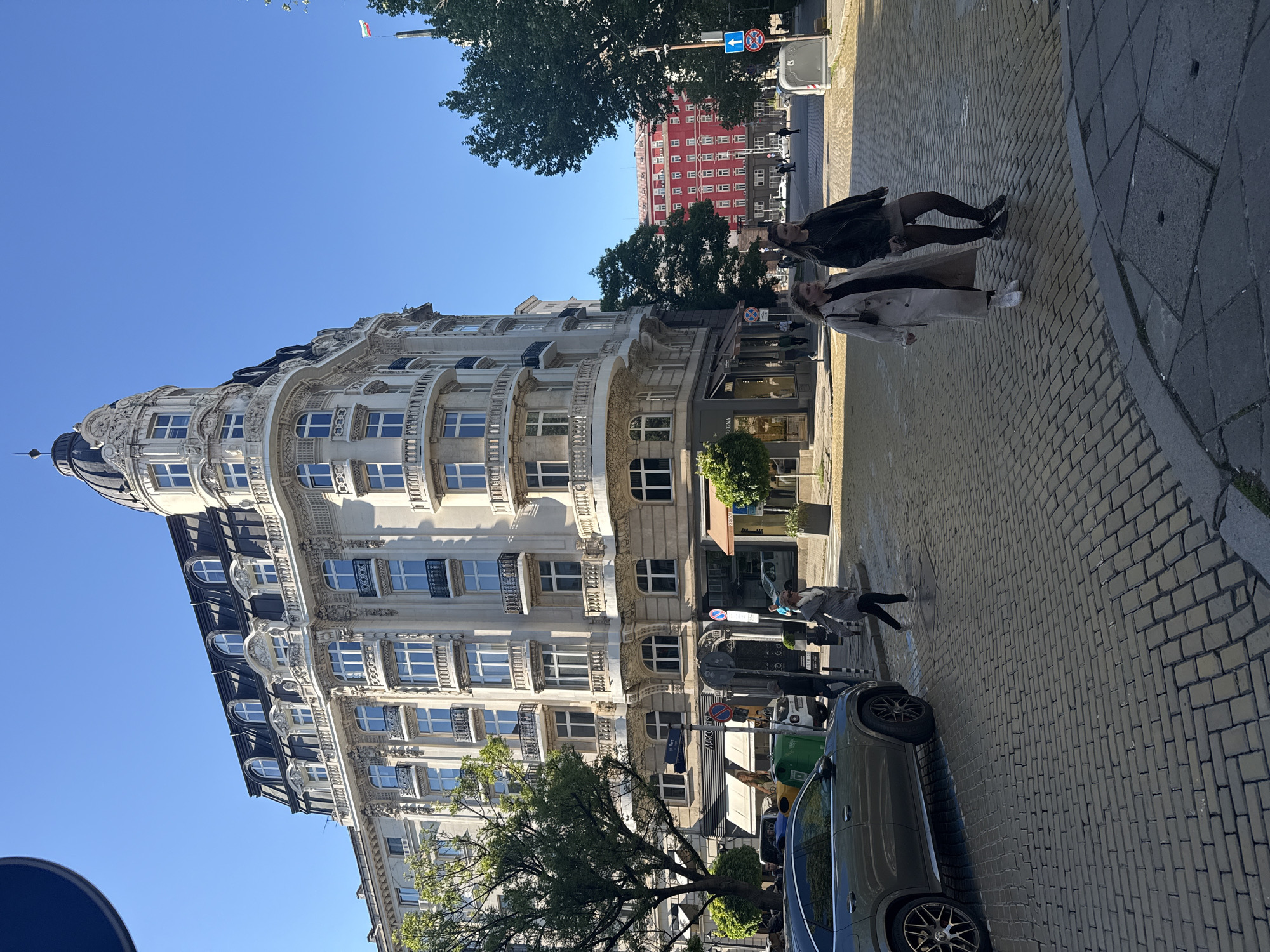 The image shows a street scene in Sofia, Bulgaria, featuring an ornate, multi-story building with a dome. The building is white with decorative architectural elements. A green car is parked along the side of the street. Pedestrians, including two women, are walking on the brick-paved sidewalk. The sky is clear blue. In the background, a red building is visible. Traffic signs are present. The street is well-lit, suggesting it is daytime.