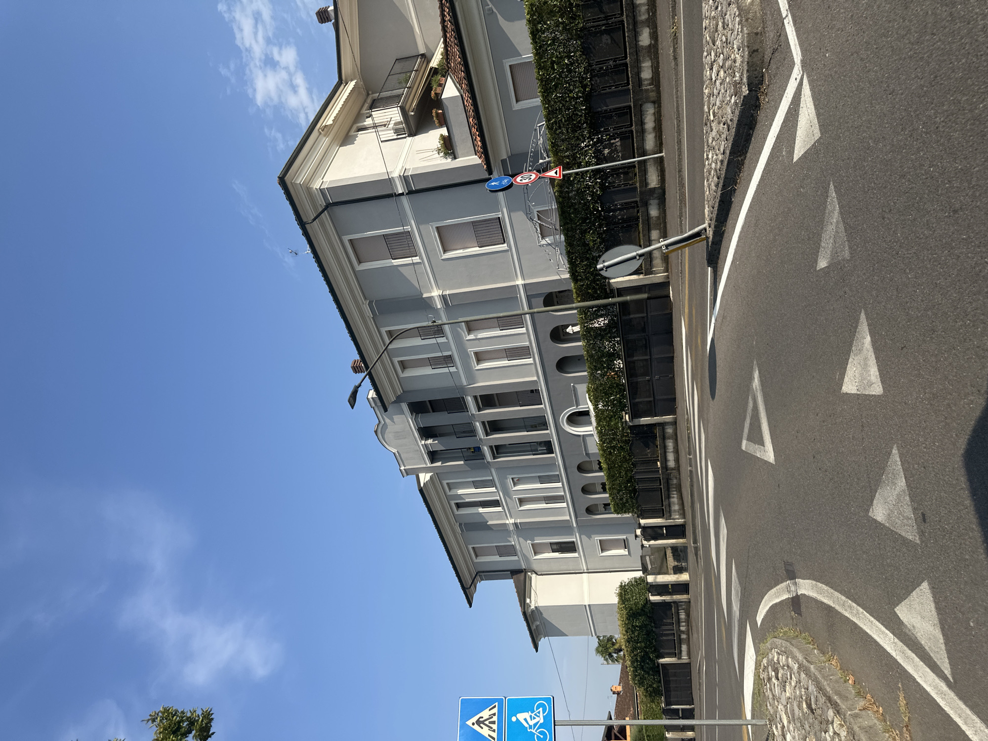 The image shows a street scene in Rudiano, Italy, on a clear day with a blue sky and scattered white clouds. In the foreground, the asphalt road curves to the left, marked with white directional arrows and a pedestrian crossing. A paved curb with cobblestones lines the edge of the road.
To the left of the road, a blue sign with a white pedestrian symbol above a white bicycle symbol is mounted on a pole. Further left, a tree with green foliage is partially visible.
Dominating the background is a large, multi-story building with a light gray facade, white trim, and dark balconies. The building has multiple windows with closed shutters. A manicured hedge forms a barrier in front of the building, behind which a dark metal fence is visible.
On the right side of the road, another pole displays two traffic signs: a circular sign with a red border and the number "30" in black, indicating a speed limit, and a triangular sign with a red border and a black symbol that appears to be an exclamation mark, warning of a hazard. A lamppost is also visible near the building. The overall scene depicts a residential or commercial area in a town.