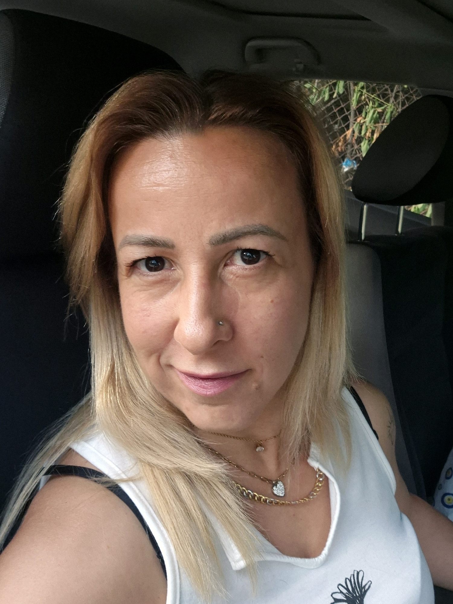 A medium shot selfie captures a woman sitting in a car. She has blonde hair with brown undertones, fair skin, and a light makeup application including pink lipstick and subtle eye makeup. She is wearing a white tank top with black straps visible underneath and two layered gold necklaces. A small nose stud is visible on her right nostril. The background shows the interior of a car with a black headrest and a portion of the seat visible. Outside the car window, greenery and a wire fence can be seen, suggesting an outdoor, possibly suburban or rural, location. The lighting appears to be natural daylight.