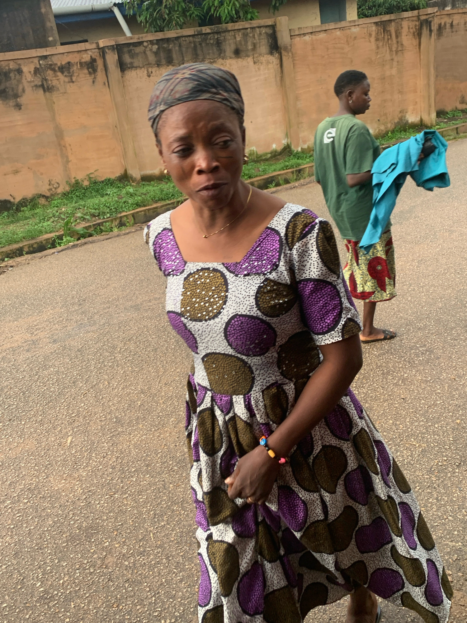 In Jos, Nigeria, two individuals are observed standing on an asphalt road adjacent to a concrete wall. The person in the foreground, a woman, is wearing a headscarf and a vibrantly patterned dress adorned with purple and brown circular motifs. She has a beaded bracelet on her left wrist. Behind her, to the right, another person, whose gender is not clearly discernible, is wearing a green t-shirt with a white logo and patterned shorts. This individual is holding a piece of blue fabric. The environment suggests an outdoor urban setting with some vegetation visible along the roadside.