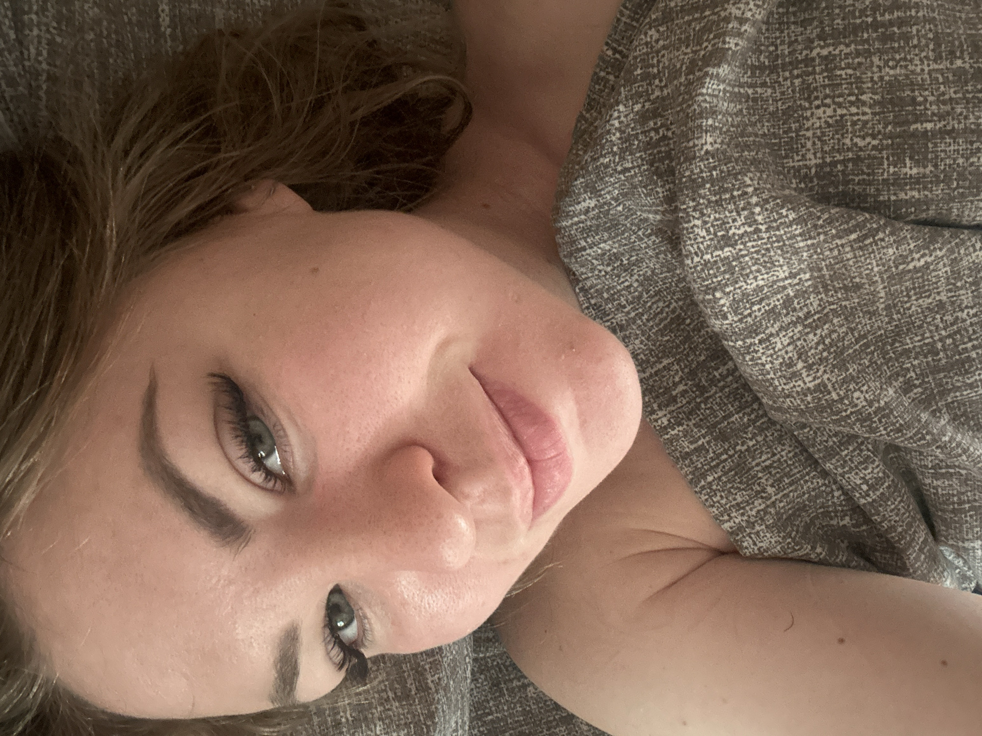 A close-up, overhead, selfie-style photo shows a fair-skinned woman lying down. She has medium-length, light brown hair, green eyes, and is wearing false eyelashes. Her eyes are looking towards the camera, and she has a slight smile on her lips, which are lightly glossed. She is wrapped in a textured grey blanket that covers her shoulders and chest. The background is a continuation of the grey textured blanket, suggesting she is in a bed. The lighting is soft, creating a warm tone. There is no discernible urban environment or location information present in the image.