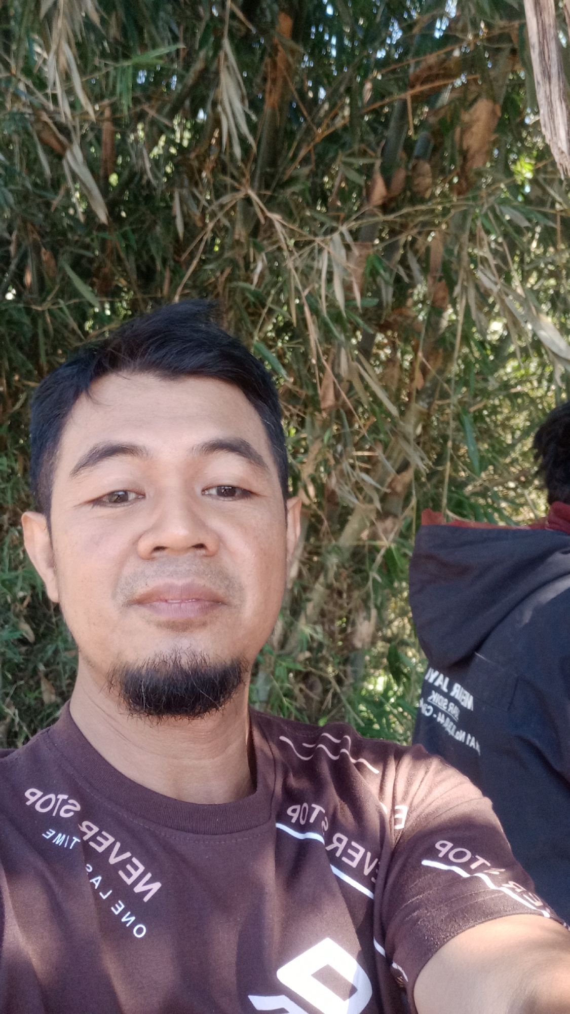 A man with a goatee and dark hair is taking a selfie outdoors. He is wearing a brown t-shirt with white lettering that appears to be in reverse. Behind him, there is lush green foliage, suggesting a natural or rural setting. To his right, another person is partially visible, wearing a black hooded jacket with white text on the back. The lighting suggests it is daytime, possibly morning or afternoon, with sunlight filtering through the leaves. The image was captured in Desa Burujul Jaya, Indonesia.