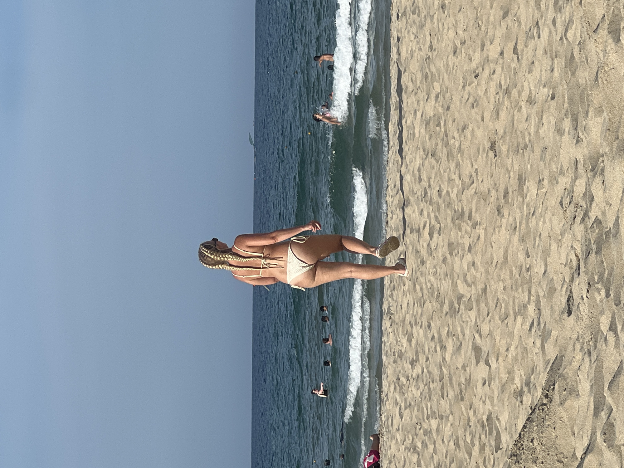 An image taken from behind a woman walking on a sandy beach towards the ocean. She has blonde hair in braids and is wearing a light-colored bikini. In the background, the ocean is calm with small waves. Several people are visible in the water, some swimming and others standing. The sky is clear and blue. This scene is consistent with a typical day at a beach in Da Nang, Vietnam, known for its popular coastal areas like My Khe Beach and Non Nuoc Beach.