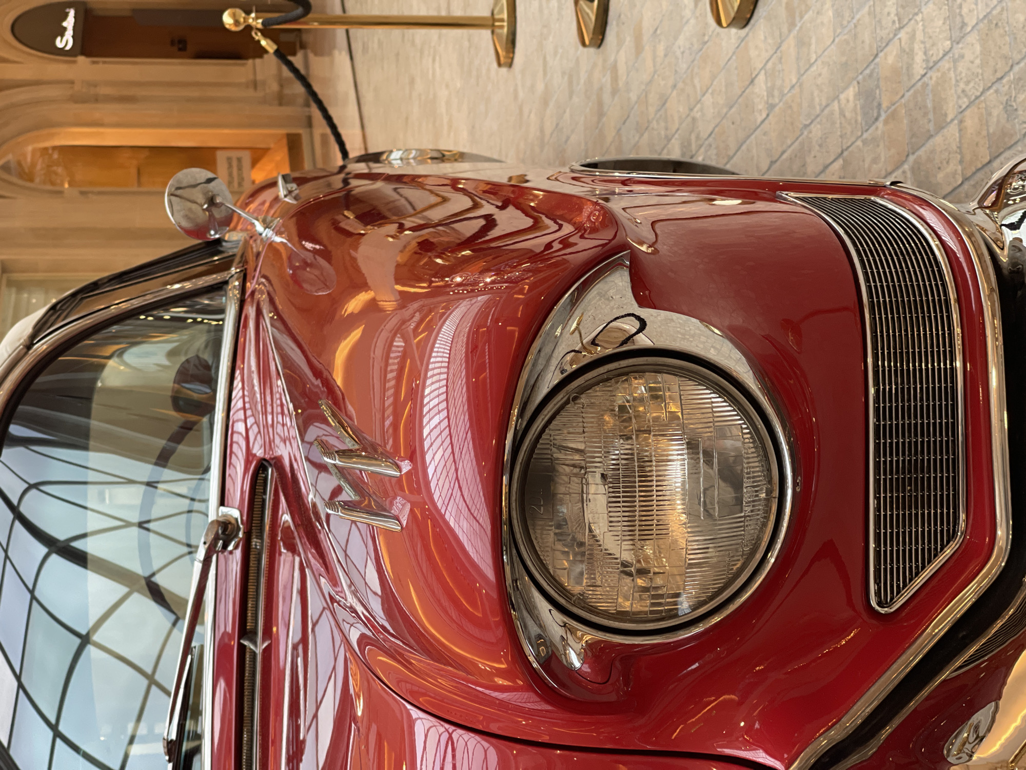 This media file is a close-up, angled shot of the front of a vintage red car. The car's shiny, polished surface reflects the surrounding environment, including what appears to be an interior setting with tiled walls and a domed ceiling or archway.
The subjects are the car itself, with its prominent round headlight, chrome accents, and a vertical grille. There are no people visible. The setting is likely an indoor exhibition space or a museum, given the polished flooring, decorative railing with a rope, and the general presentation of the car.
Notable details include the car's vibrant red paint, the intricate design of the grille, and the detailed lens of the headlight. The reflections on the car's body suggest a well-lit environment, possibly indoors with artificial lighting. The image is oriented vertically.
There is visible text on a dark sign in the upper left portion of the image. It appears to be a logo or nameplate with stylized lettering, possibly reading "Shelby."
The overall impression is one of an appreciation for automotive design and history, showcasing a classic vehicle in a refined setting. There are no explicit cues about the time of day or weather as the scene is indoors.