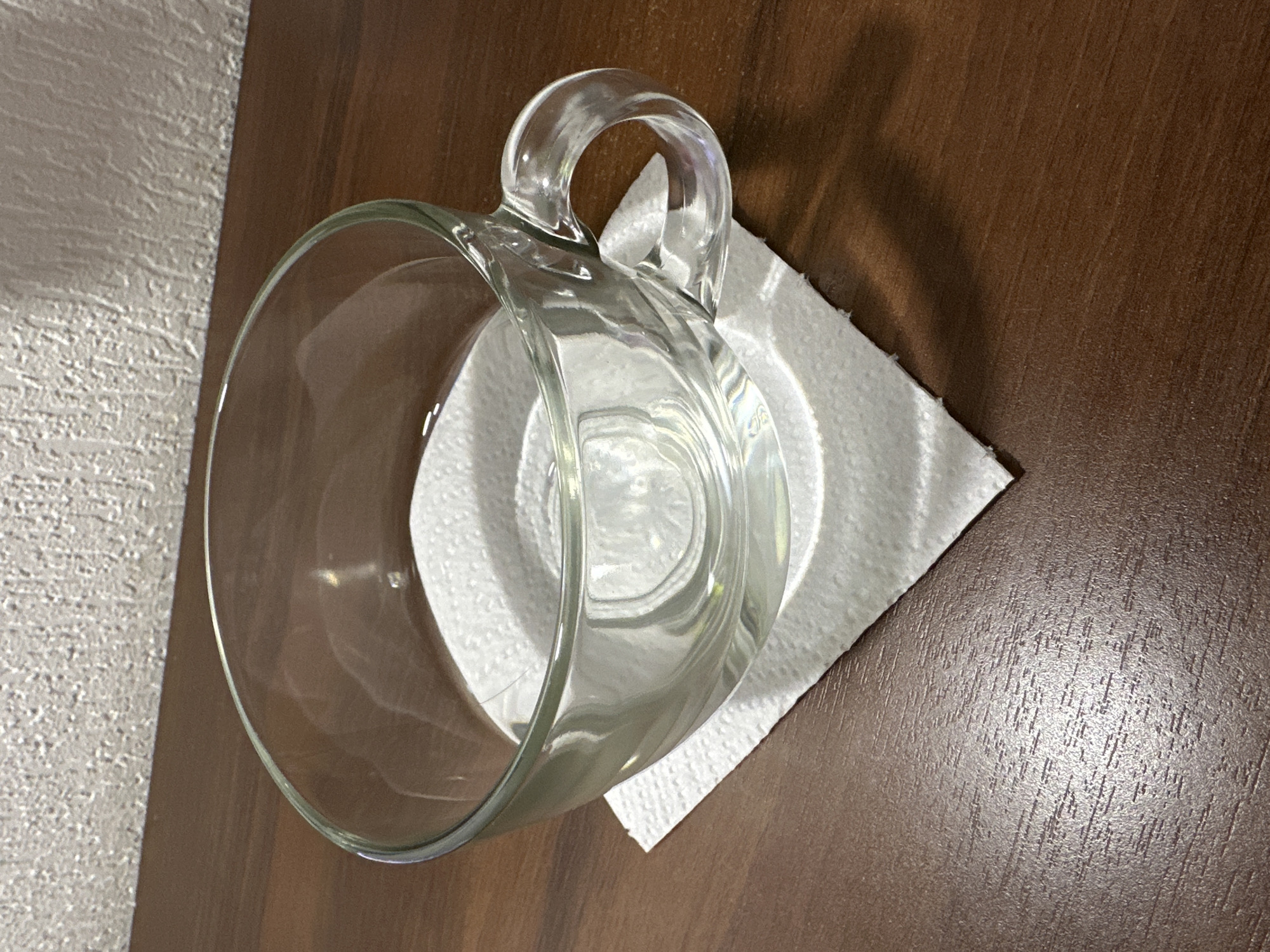 A clear glass mug filled with water sits on a white paper napkin, which is placed on a brown wooden surface. The background consists of a textured white wall. The lighting creates shadows and reflections on the objects and surfaces. The scene appears to be indoors, possibly on a table in a home or office. There is no visible indication of the city of Novosibirsk, Russia.