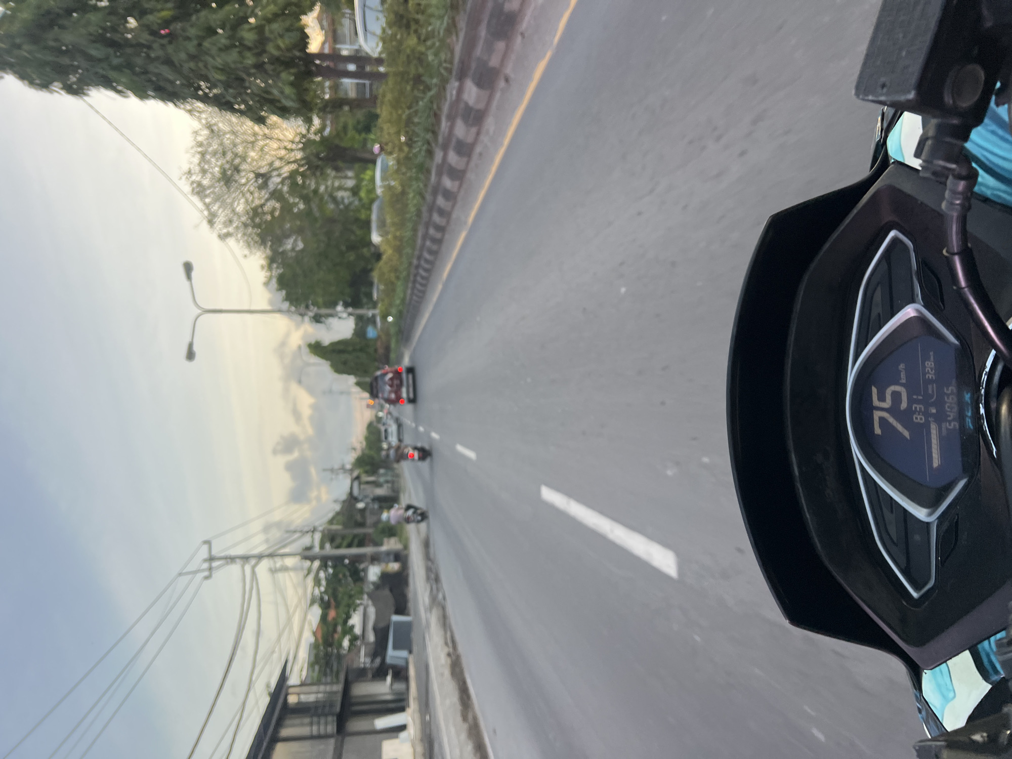 This image depicts a point-of-view shot from a motorcycle dashboard, likely traveling on a road in Denpasar, Indonesia, at dusk. The digital display on the motorcycle shows a speed of 75 km/h and a time of 8:31, with a trip meter reading of 328. The odometer shows 54065. The road ahead is paved, with white lane markings and a yellow line delineating the edge. On the left side of the road, there are buildings and power lines, indicative of an urban or suburban environment. On the right, a median with greenery and trees separates the lanes. Several vehicles, including motorcycles and cars, are visible on the road, suggesting moderate traffic. The sky is a soft gradient of blue and orange, typical of sunset.