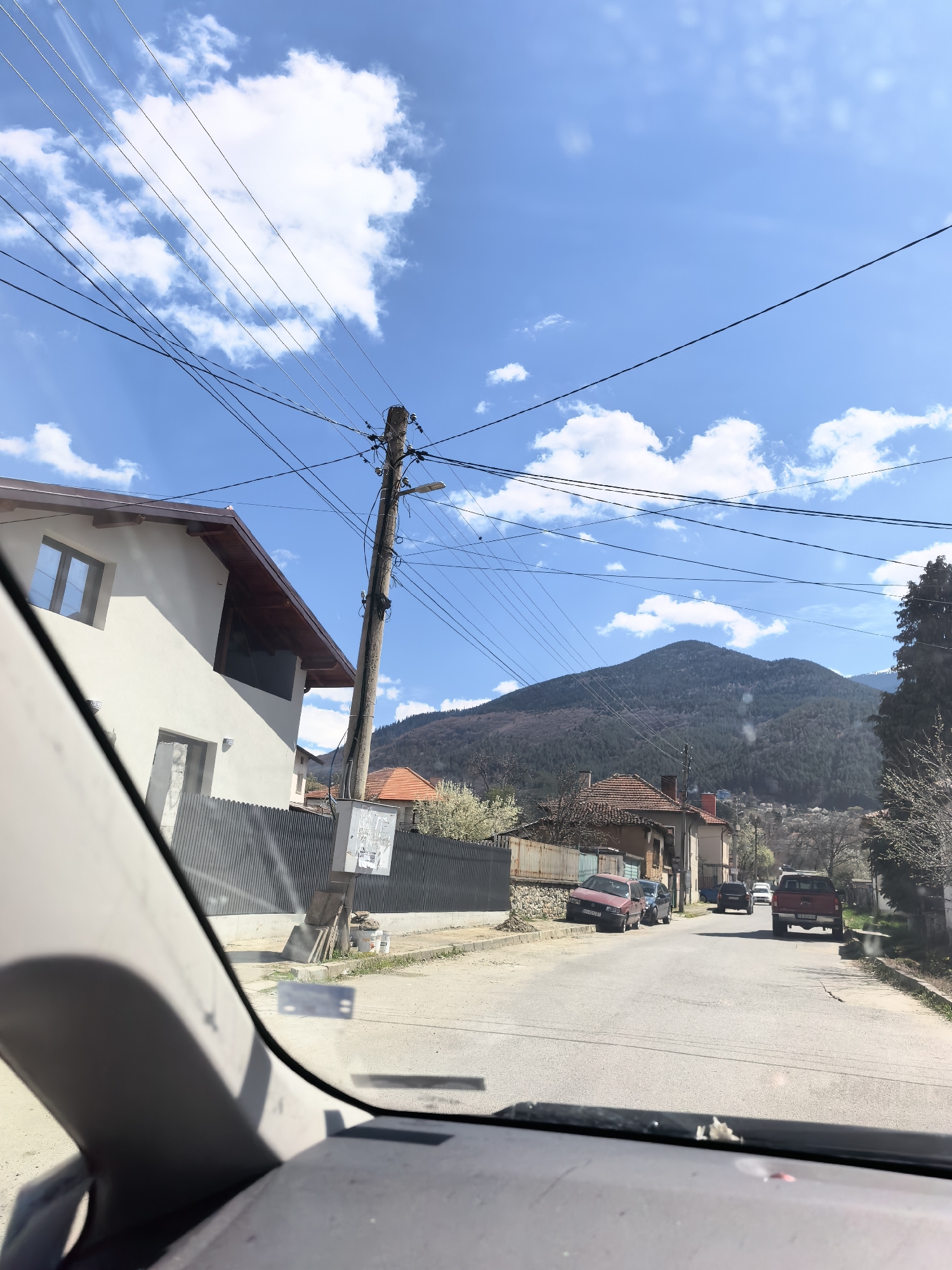 The image shows a street view from inside a vehicle. The location appears to be a residential area, possibly a small town or village, set against a backdrop of a mountain range under a partly cloudy sky. There are houses on either side of the road. On the left is a modern white house with a gray fence. Further down the road, there are older buildings with red tile roofs. Several cars are parked along the street. A red pickup truck is driving away, followed by other vehicles. Power lines are visible overhead, connecting to a utility pole near the modern white house. The image is taken from the driver's perspective, with the dashboard and part of the windshield visible in the foreground.