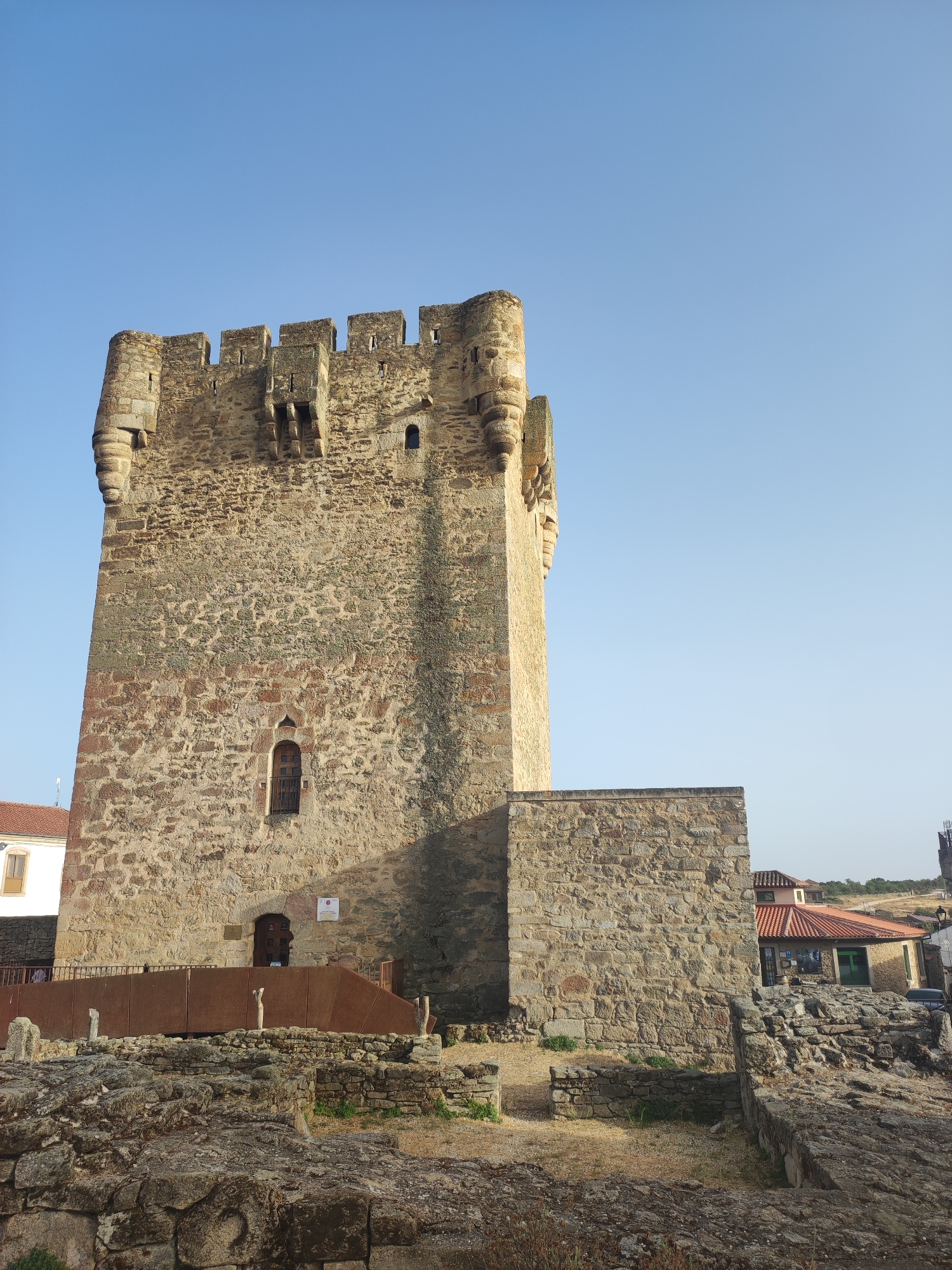 The image shows a stone tower in the city of Sobradillo, Spain. The tower is ancient, with a rough-hewn stone construction and crenelated battlements. A small, arched window is visible on the main tower, and a larger, darker wooden door is located at its base. A modern, rust-colored ramp leads up to the tower entrance, with a railing on either side. In the foreground, there are ruins of more stone structures, suggesting the tower is part of a larger, historical complex. The sky above is clear and blue. To the right of the tower, there is a building with a red tile roof, and in the distance, more buildings and a hillside are visible. No people are clearly visible in the image.
