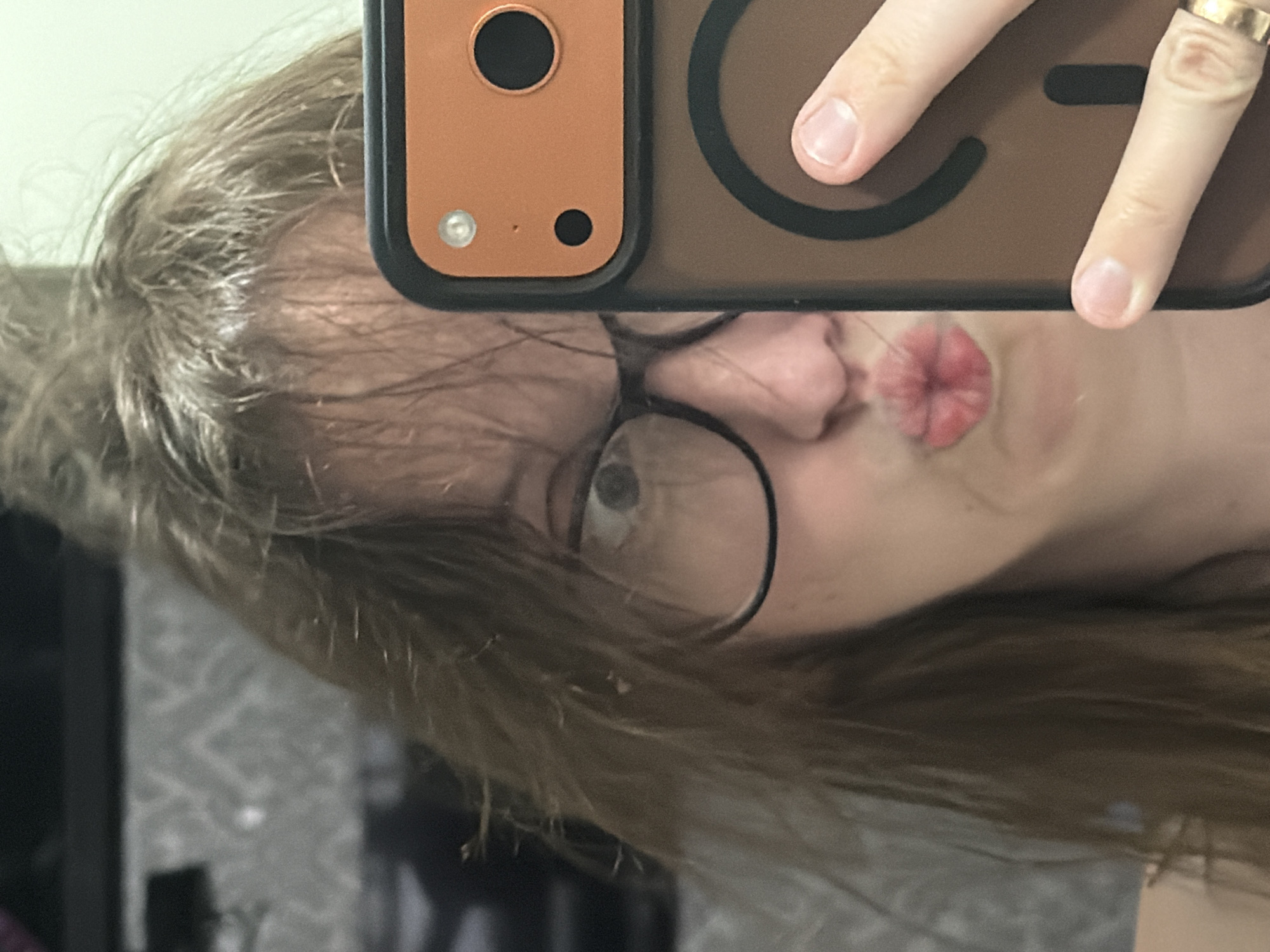 A person is taking a selfie in a mirror. They are wearing glasses and have their lips pursed. Their hair is disheveled. They are holding a phone with an orange case. The background appears to be a room with patterned wallpaper and furniture. The image does not contain any information that would allow for the determination of the location of Bila Tserkva, Ukraine.