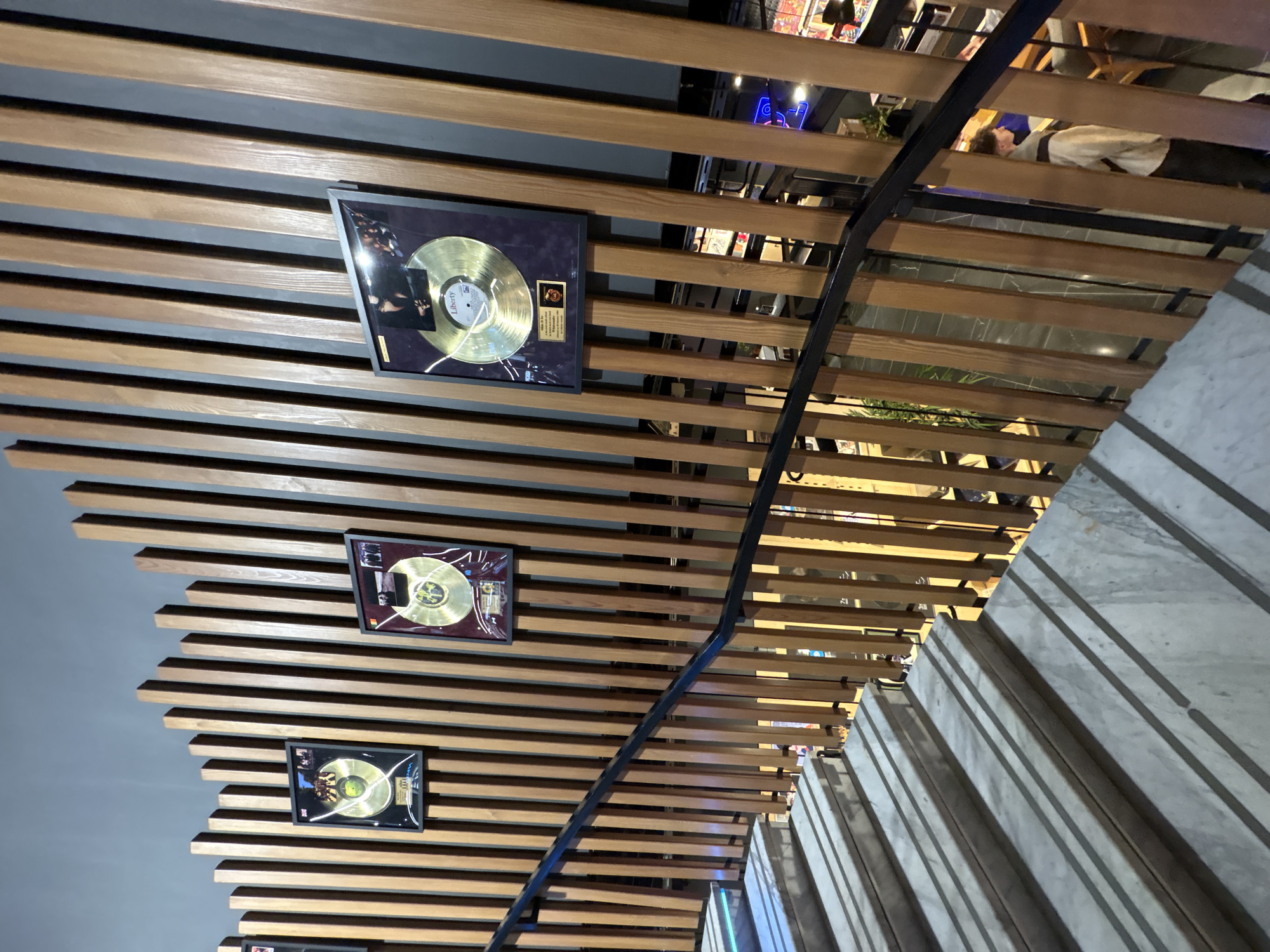 The image shows a modern interior with a slatted wooden ceiling, on which three framed gold record awards are mounted. A black staircase railing diagonally crosses the view. In the upper right, indistinct figures are partially visible through the wooden slats. To the right are grey and white steps. The scene likely depicts a restaurant or entertainment venue interior in Moscow, Russia, given the presence of gold record awards, which are often found in such establishments. The picture suggests a focus on music or entertainment.