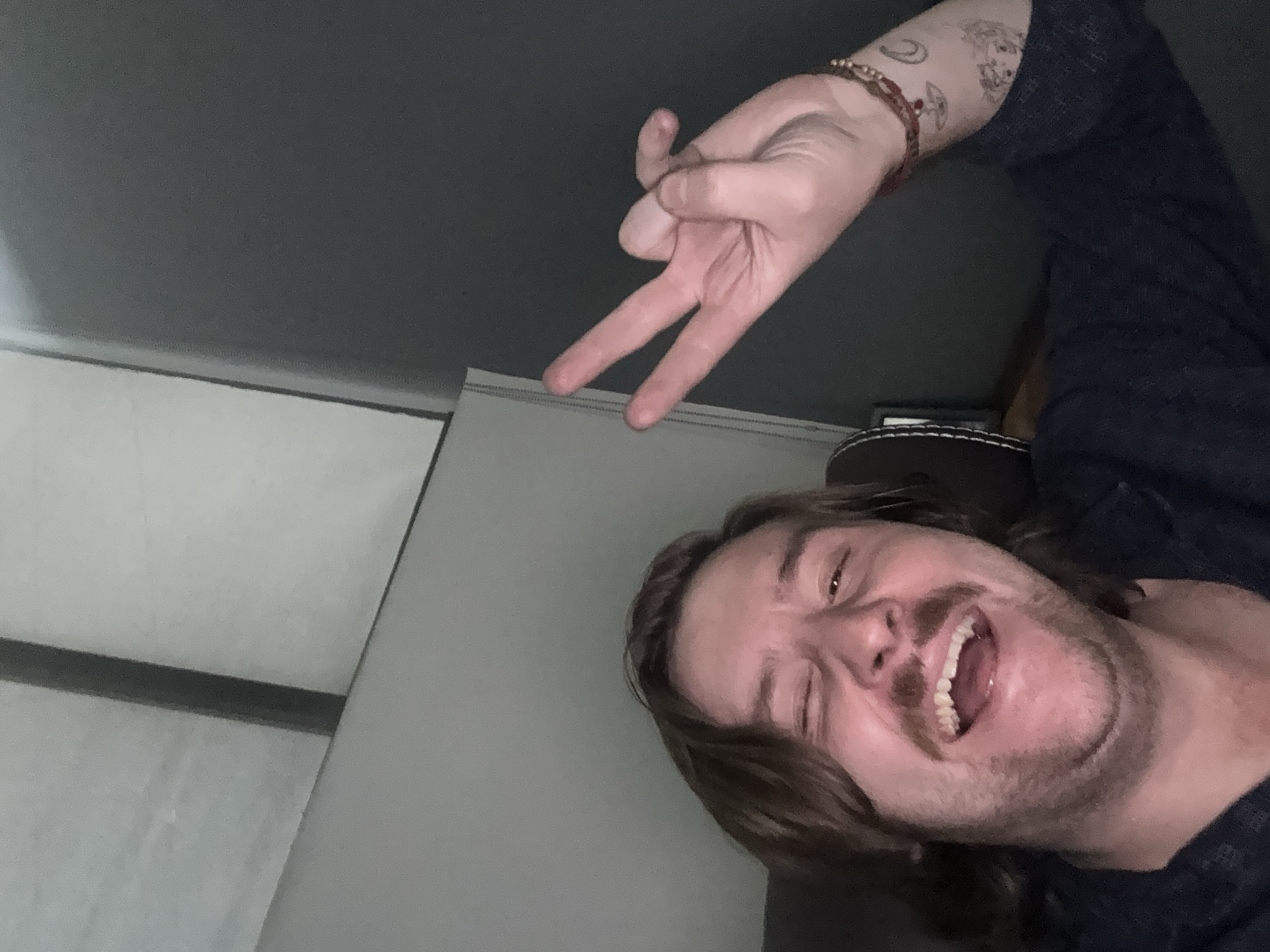 A man is lying down indoors, possibly on a couch or bed. He is smiling and winking. His right hand is raised, displaying a "peace sign." The man has long brown hair and a mustache. A bracelet adorns his wrist, and there is a tattoo on his forearm. The background includes a grey wall, a grey ceiling, and a white screen or partition. The location is difficult to determine, but there is no information indicating it is in Cuautitlán Izcalli, Mexico. There is not enough information to confirm the location.