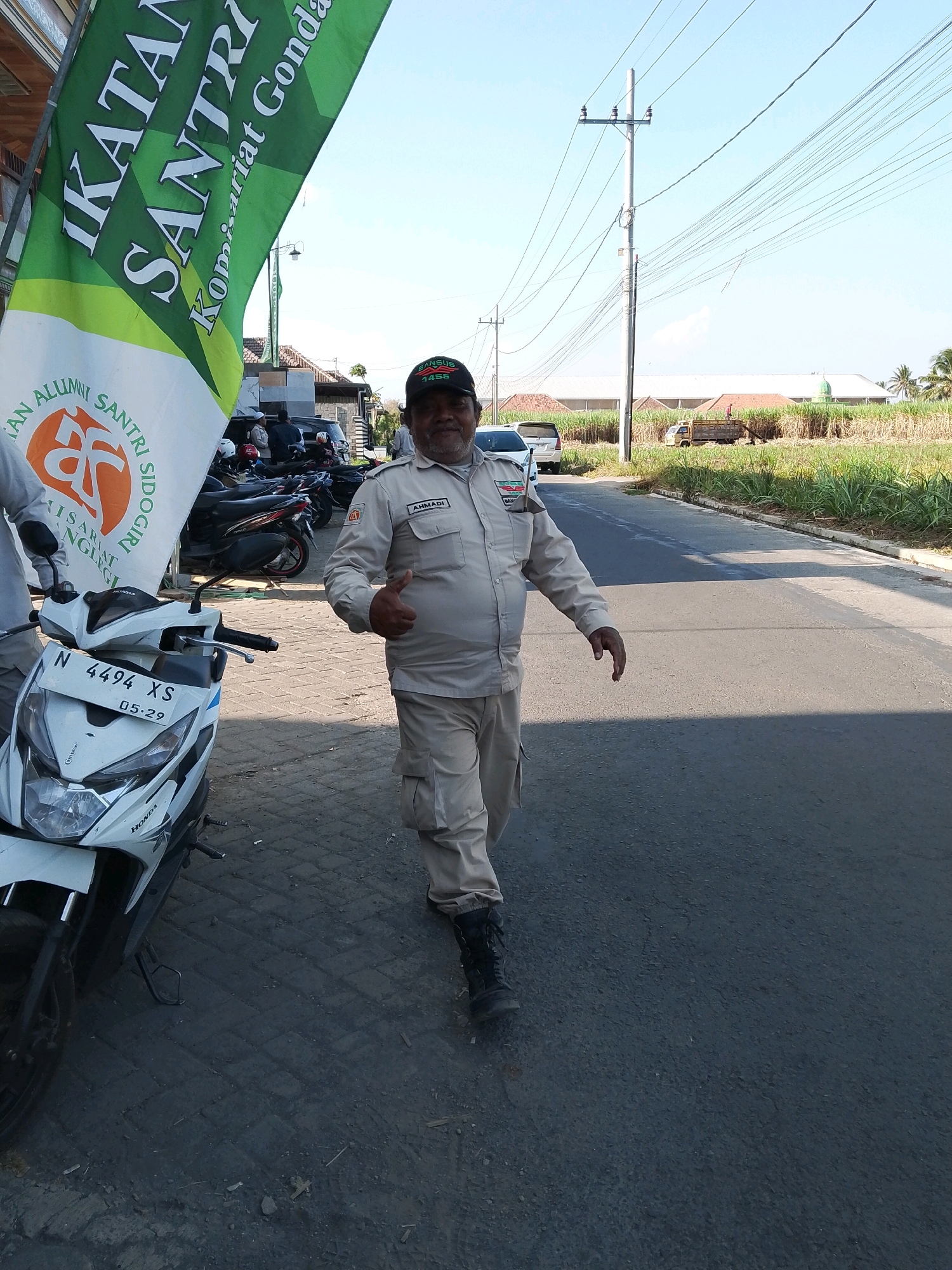 In Banjarejo, Indonesia, a man in a light brown uniform and cap stands on the side of a road. He is giving a thumbs-up gesture. A white scooter is parked to his left, and a green banner is visible behind him. In the background, there are buildings, sugarcane fields, a truck, and utility poles. The sky is clear and blue.