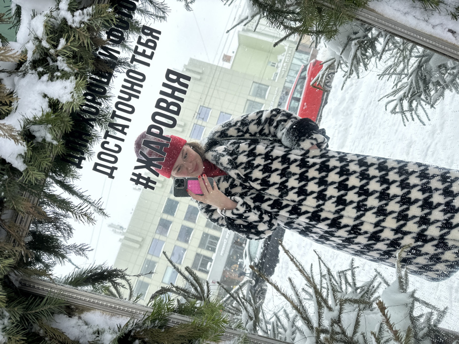 A female individual is depicted taking a mirror selfie in an urban, snowy environment in Kazan, Russia. She is wearing a red knit beanie, glasses, a red turtleneck, and a black and white houndstooth pattern faux fur coat. Her right hand holds a dark-colored smartphone with a vibrant pink case, positioned to capture her reflection. Her fingernails are painted red. The mirror is vertically oriented and framed by snow-laden evergreen branches, with additional snow visible on the branches and the ground in the reflection.
The background, reflected in the mirror, shows a snow-covered street with a red compact SUV or hatchback partially visible, along with other vehicles and multi-story buildings under an overcast sky with active snowfall. Russian text is affixed directly onto the mirror surface, reading "ДЛЯ ХОРОШ И ФОТ ДОСТАТОЧНО ТЕБЯ #ЖАРОВНЯ". The hashtag "#ЖАРОВНЯ" (Zharovnya), which translates to "brazier" or "roaster," is a known restaurant chain, indicating the scene may be at or near a Zharovnya establishment in Kazan.