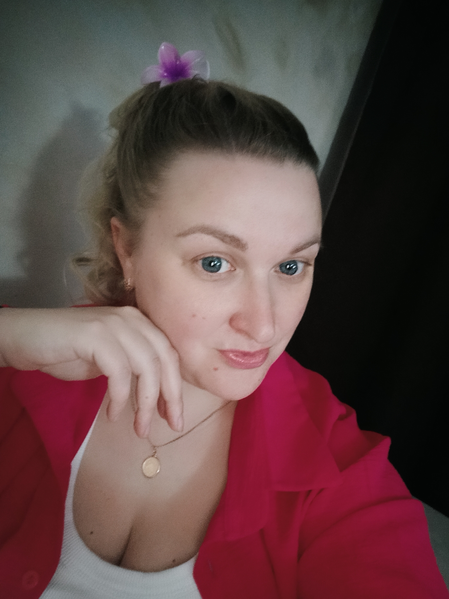 An indoor, close-up portrait of a woman, likely a selfie, taken in Chaikovsky, Russia. The woman, an adult with blue eyes, is positioned slightly off-center to the right, looking directly at the camera with a subtle, pleasant expression and a slight smile. She is seen leaning her chin on her right hand, with her fingers gently cupping her jawline.
Her light brown or dirty blonde hair is styled in a partial updo, featuring a translucent purple flower-shaped hair clip or tie at the crown of her head. She wears a bright pink or magenta collared shirt or jacket over a white ribbed tank top or camisole, a portion of which is visible, showing a hint of cleavage. A delicate gold-colored chain necklace with a small, round gold pendant rests on her chest.
The background consists of a textured, light grey or beige wall on the left side, showing some shadow details, and a dark, indistinct vertical surface on the right. The lighting is soft and even, highlighting her facial features. There is no visible text in the image.