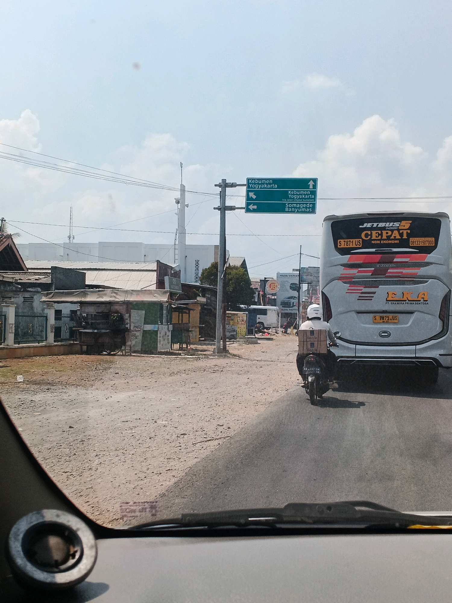 The image depicts a street scene in Kedungpring, Indonesia, on a clear, sunny day. A large white tour bus with "JETBUS CEPAT" and "EKA" branding is the central focus, traveling on an asphalt road. The bus has license plate "S 7871 US". In front of the bus, a motorcycle rider wearing a white helmet and carrying several brown boxes on their back is also traveling in the same direction. On the left side of the road, a row of small, rustic buildings with corrugated metal roofs and weathered walls line a dirt path. A green road signpost with white lettering indicates directions to "Kebumen Yogyakarta" and "Somagede Banyumas". Power lines are visible overhead, strung between utility poles. The environment is characterized by a mix of urban and rural elements, with a bright, overcast sky.