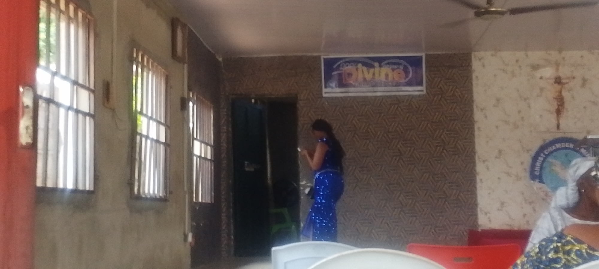 The image captures an interior scene in Lokoja, Nigeria. A woman in a bright blue, sequined dress stands near a doorway. Above her, a sign reads "Cool Divine World Ministry." The wall behind her features a patterned design. To the right, a crucifix hangs on a different wall, along with a circular sign that reads "Christ Chamber." The foreground features white and red chairs, suggesting a gathering place. Windows with metal bars are visible on the left side of the image. The walls around the windows appear to be concrete. A ceiling fan is present.