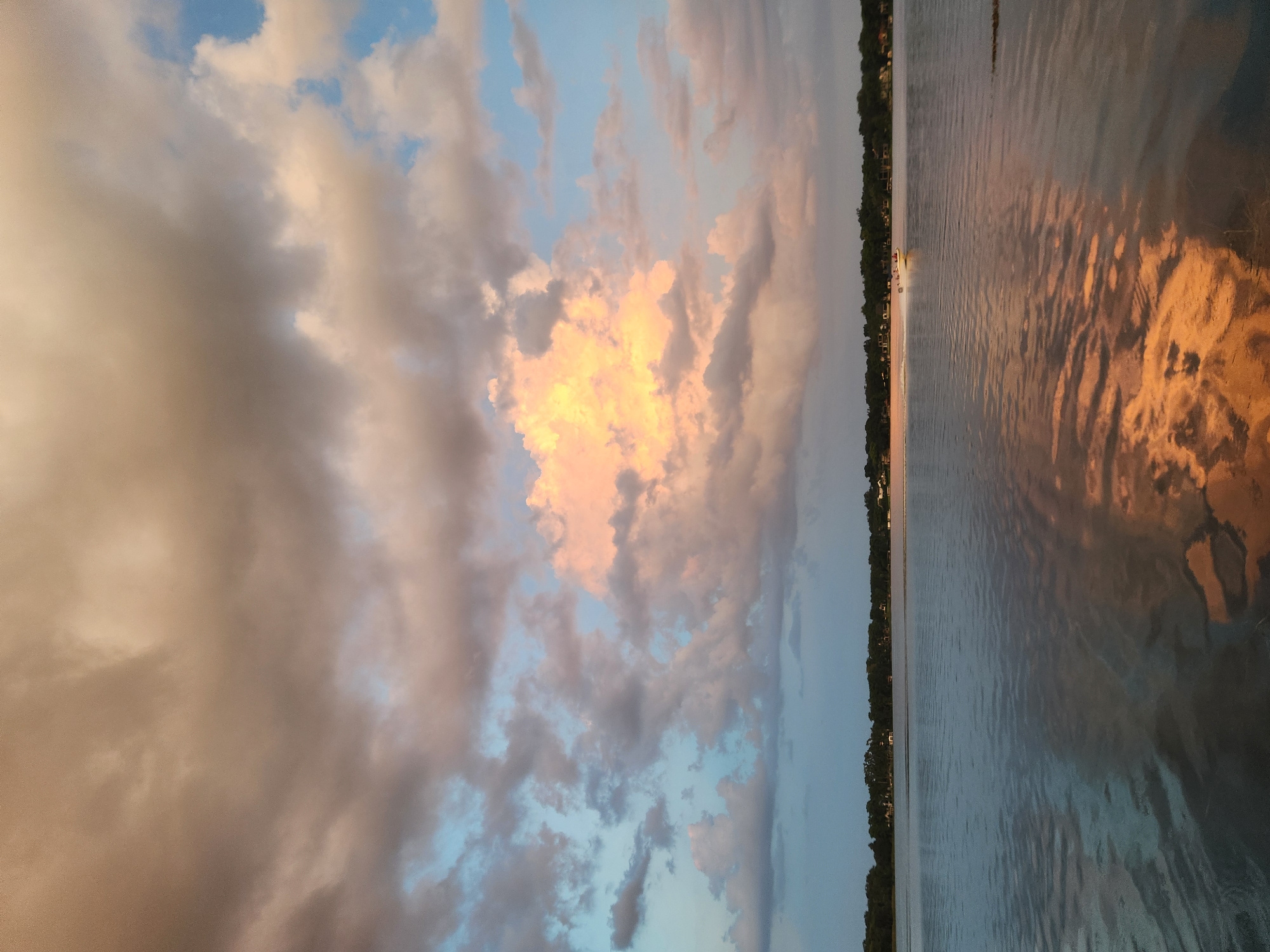 The image depicts a serene lake scene in Winter Park, United States of America, under a dramatic sky filled with colorful clouds. The sky is a canvas of soft blues, pinks, and oranges, with a prominent large cloud illuminated by sunlight, creating a warm glow. The water's surface reflects the sky's hues, adding to the tranquil ambiance. Along the horizon, a line of trees and scattered houses suggest a suburban or natural setting. A small white boat with people aboard is visible on the right side of the lake, leaving a wake as it moves. The overall impression is one of a peaceful evening at dusk.