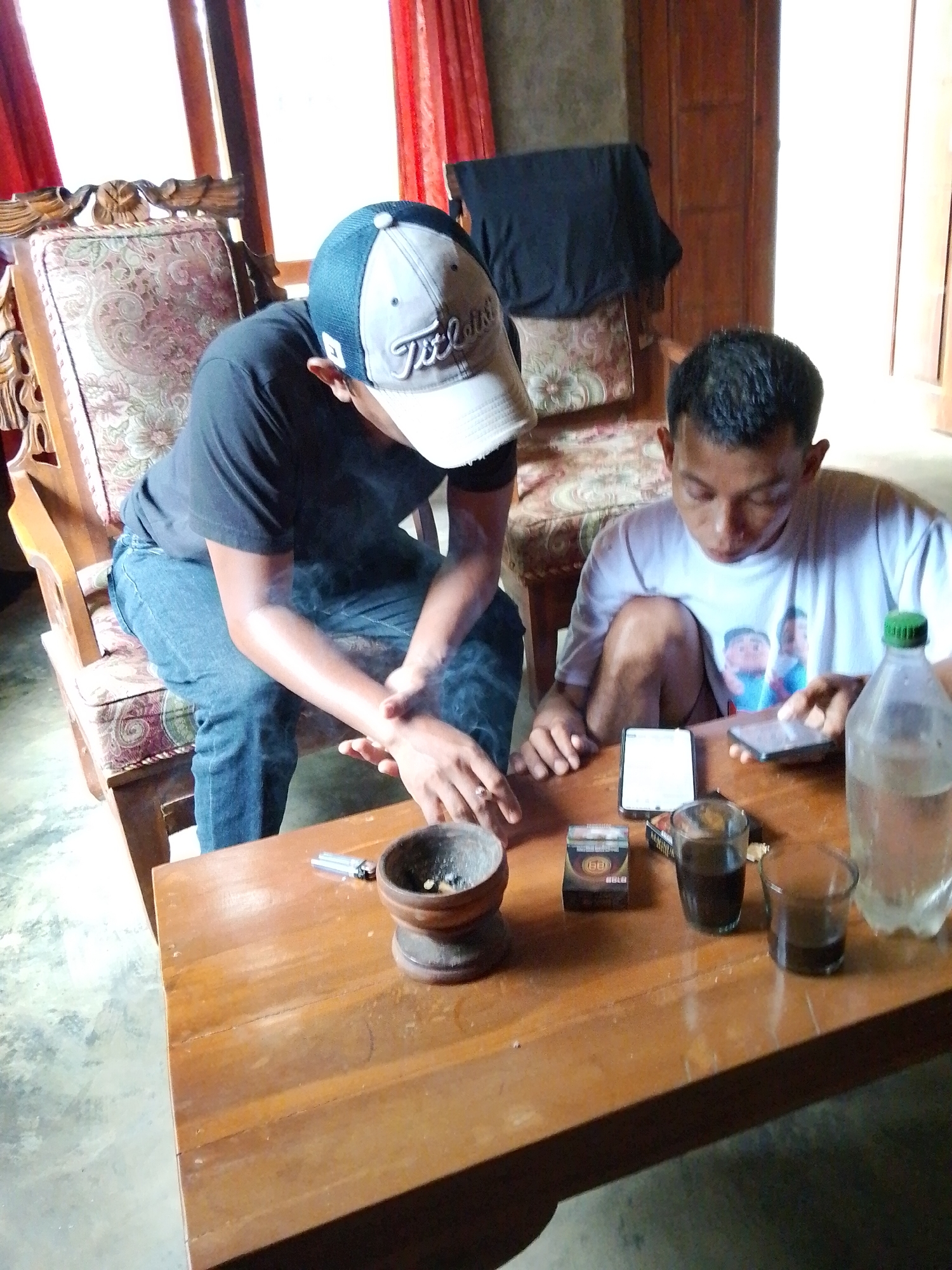 Two young men are sitting at a wooden table in a room that appears to be in Madukoro, Indonesia. One man, wearing a black t-shirt and a baseball cap, is sitting on a patterned armchair. He is smoking, with visible smoke rising from his hands. The other man, wearing a white t-shirt, is sitting on the floor, looking at his phone. On the table, there are two small glasses containing a dark liquid, a pack of cigarettes, two smartphones, a lighter, and a small ashtray. A plastic bottle of water with a green cap is also on the table. The room has red curtains in the background and wooden furniture. The overall impression is of a casual, indoor gathering.