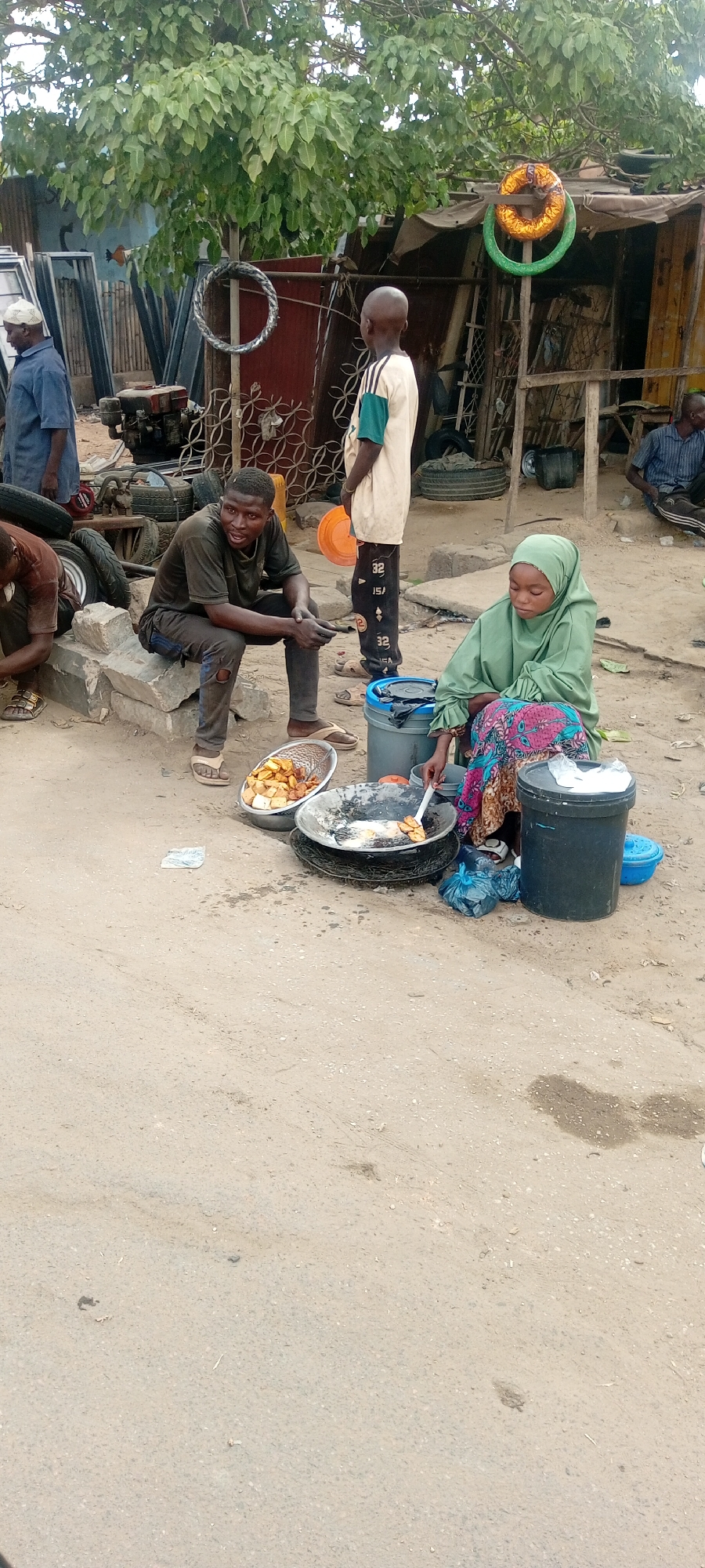 The image captures a street scene in Kano, Nigeria, where a woman wearing a green hijab and colorful patterned skirt is frying food in a large pan, likely akara (bean cakes), over a makeshift stove on the ground. A man sits on a nearby stone, holding a toothpick, with a bowl of fried food next to him. In the background, a child stands wearing a cream shirt and pants. Other people are visible in the scene including a man in blue and a man sitting off to the right. Surrounding the scene are elements such as tires, metal structures, and a variety of items, suggesting an informal outdoor setting or market area. The ground is dusty and unpaved, further indicating the urban environment of Kano.