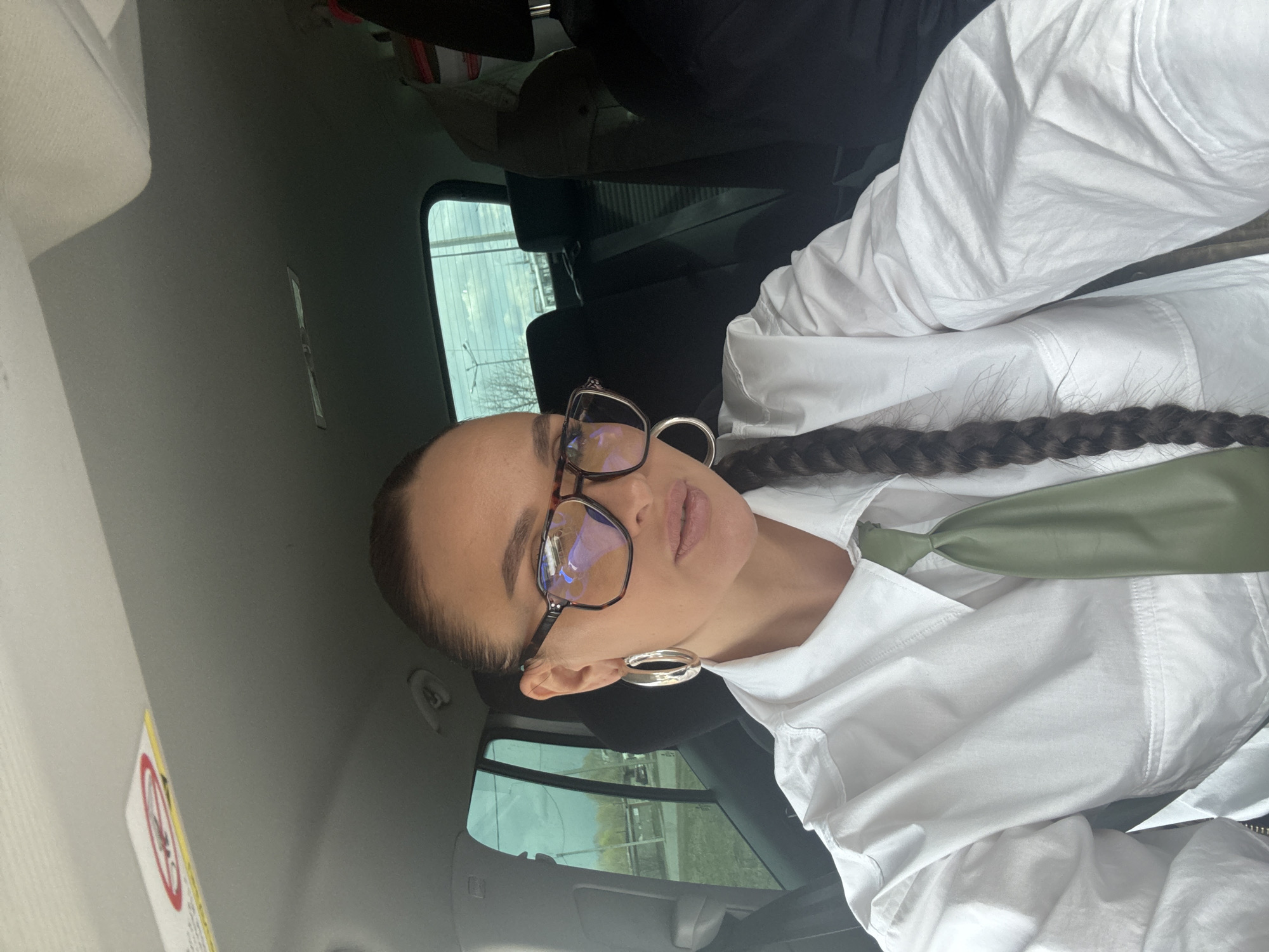 The image displays a woman seated inside a car, captured in what appears to be a selfie. She is wearing a crisp white collared shirt accessorized with a smooth, sage green tie. Her dark hair is styled in a long, neat braid that rests over her left shoulder. She wears dark-rimmed eyeglasses, likely tortoiseshell, which reflect the ambient light. Large, reflective silver-toned hoop earrings adorn her ears. Her gaze is directed towards the camera with a neutral expression.
The setting is clearly the interior of a vehicle, with dark car seats or headrests visible behind her and the car's ceiling above. Through the car window in the background, a daytime outdoor scene is visible. The sky appears overcast or lightly cloudy, with no direct sunlight. The exterior view shows faint outlines of structures and what seem to be bare trees, indicative of an urban or suburban environment, consistent with the provided location context of Chaikovsky, Russia. On the car's ceiling, a small, rectangular sticker with a red circular symbol, resembling a "no entry" or warning sign, is visible, though the accompanying text is illegible. The overall lighting suggests it is daytime, without harsh shadows.