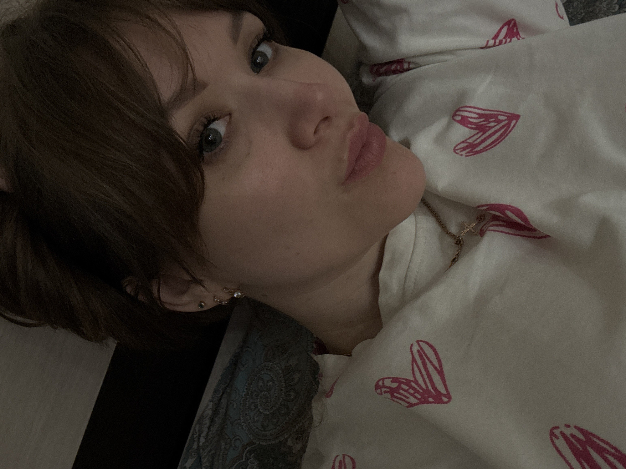 A woman is lying down, with her head turned towards the camera. She has short brown hair and light eyes. She is pouting her lips. She is wearing earrings and a gold necklace with a cross pendant. She is covered with a white blanket with pink heart designs. The location is unknown, and there is no indication that the image was taken in Saratov, Russia. The environment appears to be indoors. There are no identifiable objects indicating a specific location.