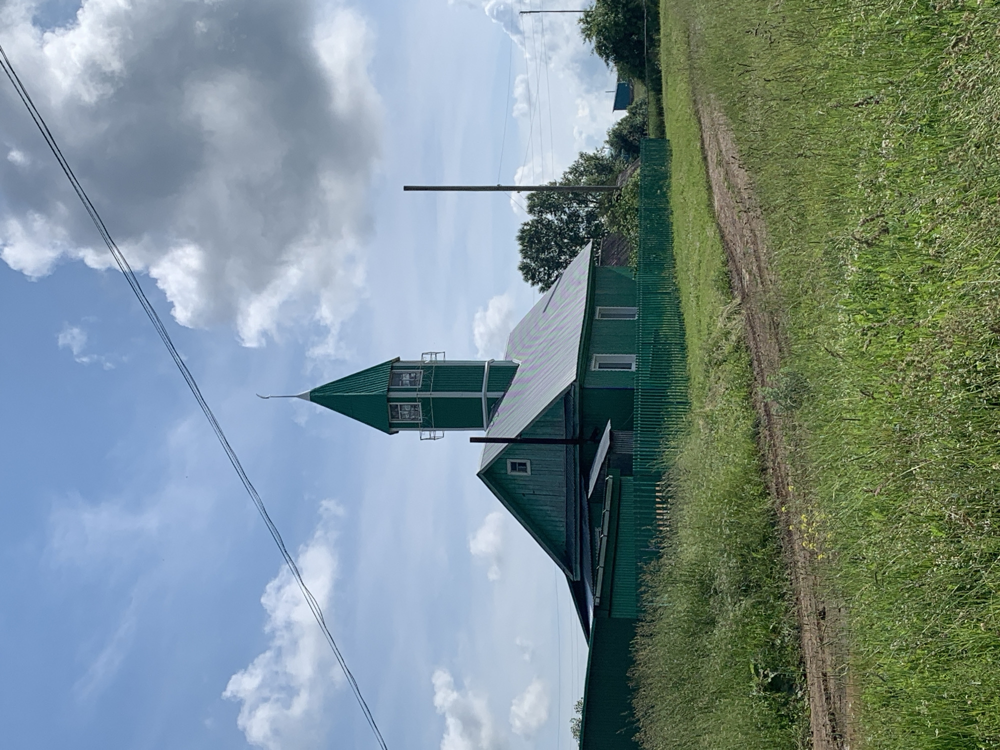 The image shows a bright green wooden building with a tall, narrow tower topped with a crescent moon and star, resembling a minaret. The building has a sloped roof and several windows. It is surrounded by a green wooden fence and tall grass. A dirt path leads through the grassy area in front of the building. The sky is blue with scattered white clouds. The building is located in the village of Barsai, Russia.