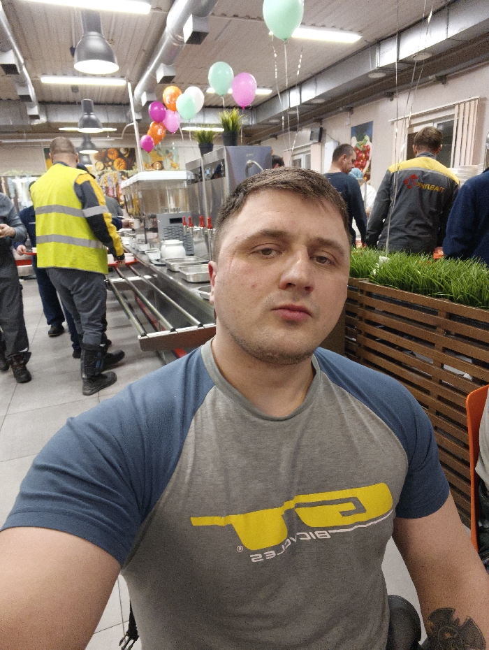 The scene is an indoor cafeteria or self-service dining facility located in Im. Gastello, Russia. In the immediate foreground, a male individual with short dark hair and a neutral expression is capturing a selfie, looking directly into the camera. He wears a grey t-shirt with blue raglan sleeves and a yellow stylized graphic logo on the chest; a tattoo is visible on his left forearm.
Behind the primary subject, a long, stainless steel food service counter is visible, featuring multiple food pans, beverage dispensers, and a tray rail. Several individuals are present in the mid-ground and background. To the left, a male wearing a yellow high-visibility vest and dark boots stands with his back to the camera at the counter. Further to the right, another male in a grey and yellow work jacket is partially visible, also with his back to the camera. Other indistinct figures are present throughout the background, engaged in what appears to be a queue or movement within the dining area.
The environment includes overhead industrial-style lighting fixtures, exposed ceiling ductwork, and light-colored tiled flooring. A cluster of balloons in shades of light green, pink, orange, and white floats above the counter area. Decorative elements comprise wall art depicting fruit imagery and a wooden planter box containing artificial green grass positioned on the right side of the frame.