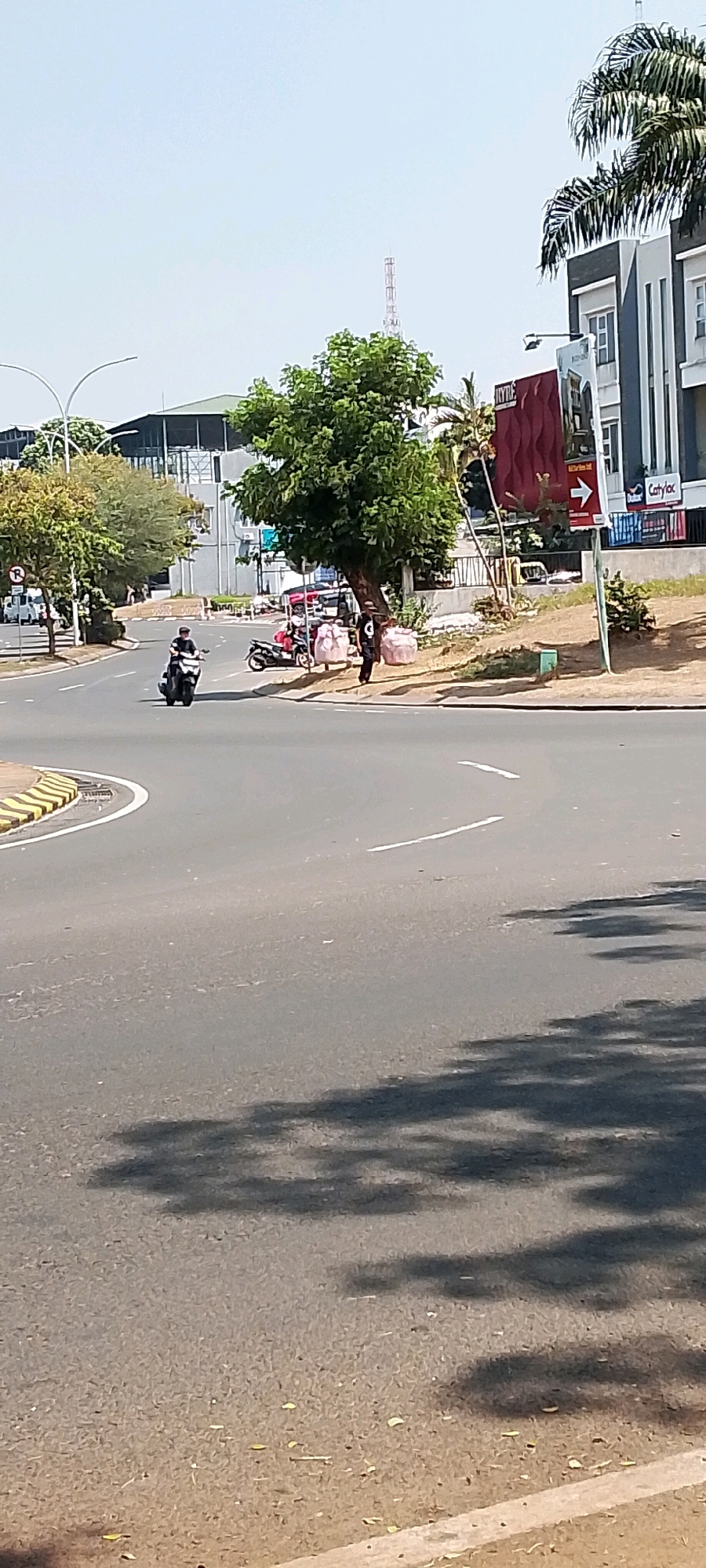 The image depicts an outdoor scene on a bright, sunny day in Gunung Putri, Indonesia. A paved road with lane markings curves through the frame, casting shadows from unseen trees onto its surface. On the left side of the road, a yellow and black striped curb is visible. A person is riding a white motorcycle down the road, heading towards the viewer. Further down the road on the right side, a red and white sign with an arrow pointing right is mounted on a pole, with a building in the background. A person in dark clothing is standing near a tree on the side of the road, holding two large white bags. Another person is seated on a red motorcycle nearby, with another red motorcycle parked beside it. Several trees with green foliage are scattered throughout the scene, and a tall transmission tower is visible in the distance. The overall environment appears to be a commercial or urban area.