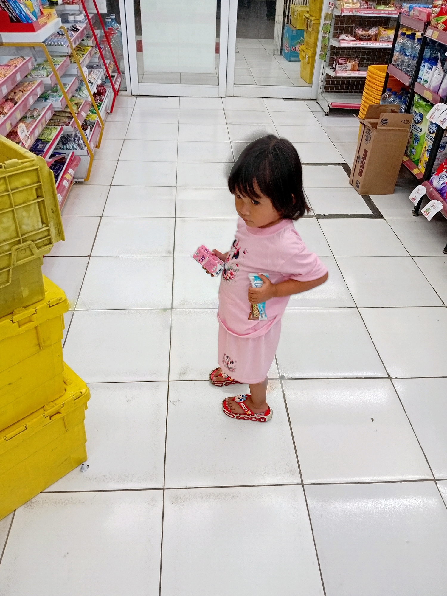 The image captures a full-body shot of a young girl standing in a brightly lit aisle of a convenience store. She is wearing a pink t-shirt and shorts set, adorned with cartoon characters. Her feet are clad in red flip-flops decorated with similar characters. In her hands, she holds a small pink box and a blue and white packaged item. The store's floor is tiled with white squares. To her left, stacked yellow crates are visible. In the background, shelves stocked with various products line the aisle, and a glass door is part of the store's entrance. The environment suggests a retail setting, possibly a convenience store in Seren, Indonesia, given the context.