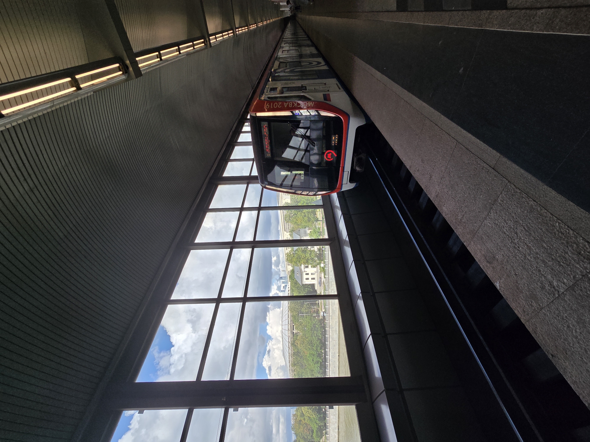 A modern red and white metro train with "MOSKVA 2019" text is stopped at a station with large windows offering a view of the Moscow River and the Luzhniki Stadium. The station features a corrugated metal ceiling with fluorescent lighting and a platform made of stone tiles. The sky is cloudy, with some patches of blue. The train's front displays a metro logo and destination information. The scene is serene and captures a moment of urban transit against a backdrop of a landmark Moscow venue.