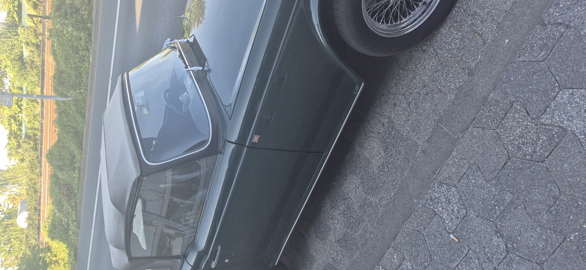 The image shows a dark green convertible sports car parked on a cobblestone sidewalk next to a street. The car has a grey fabric top, a Union Jack flag emblem on the fender, and chrome wire wheels. A discarded cigarette butt is visible on the sidewalk near the car. In the background, there is a road with a grassy embankment and trees. A street sign with German text is also visible. The scene appears to be a street in Mainz, Germany.